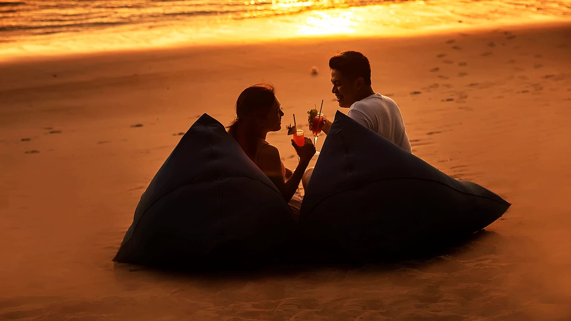 Silhouette of a couple sitting on beach beanbags enjoying tropical cocktails at sunset.
