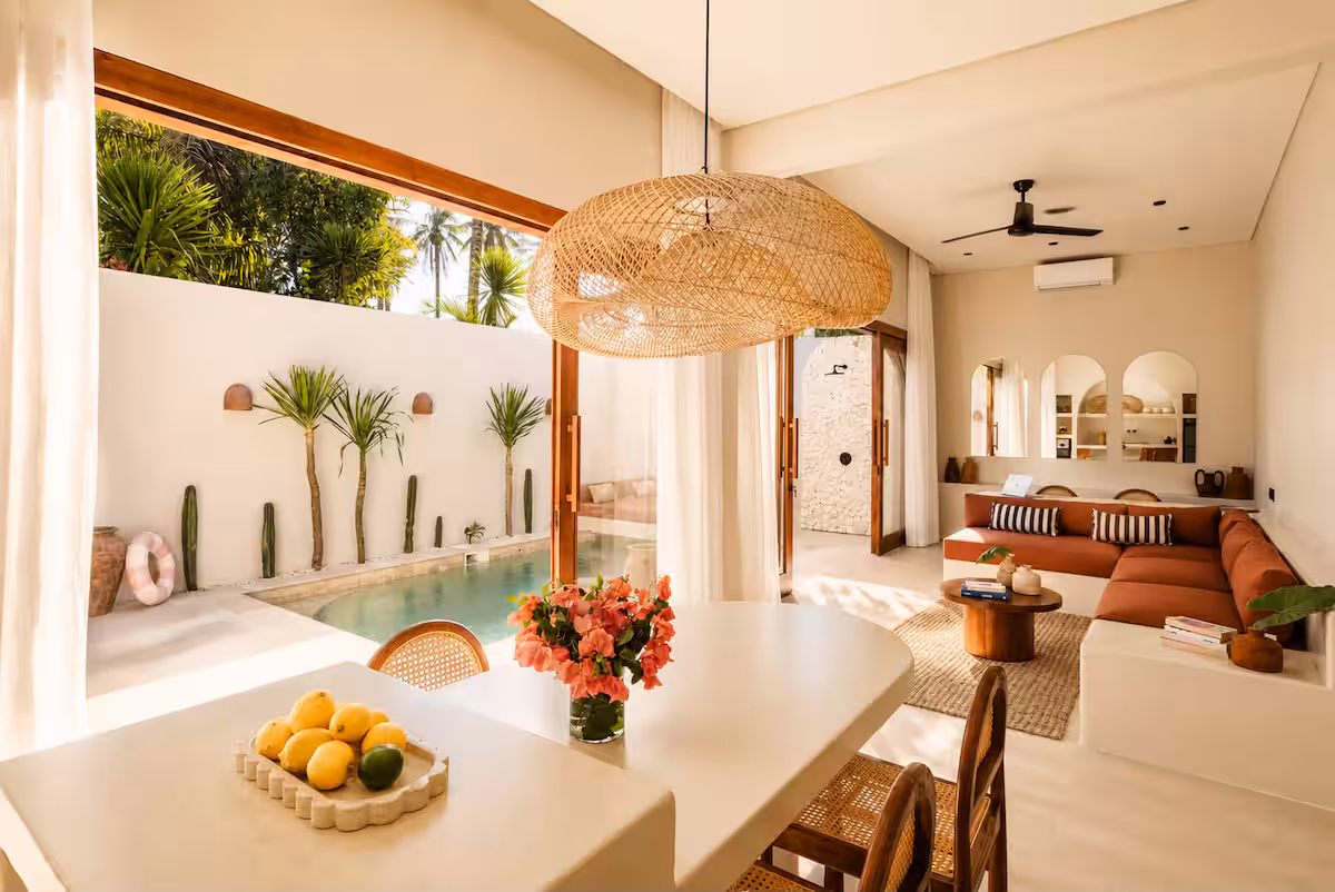 Sunlit indoor-outdoor villa interior showing a dining table with fresh flowers, a sectional couch, and wide glass doors opening to a pool.