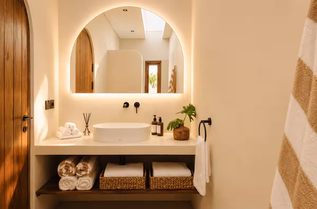 Bathroom vanity featuring an arched illuminated mirror, a vessel sink, matte black hardware, and open shelves with towels and baskets.