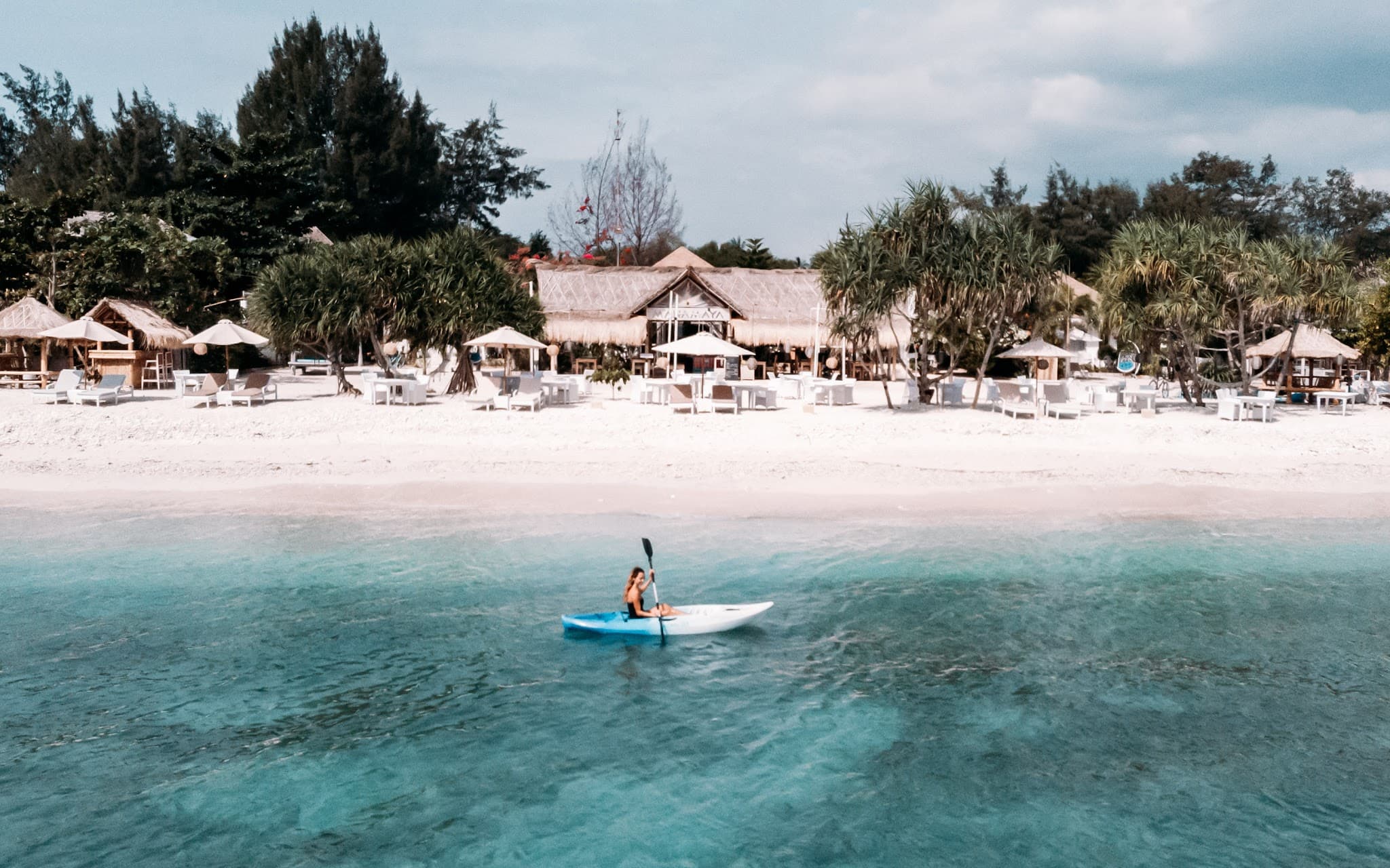 A woman kayaking in clear turquoise ocean water along a beachfront resort.
