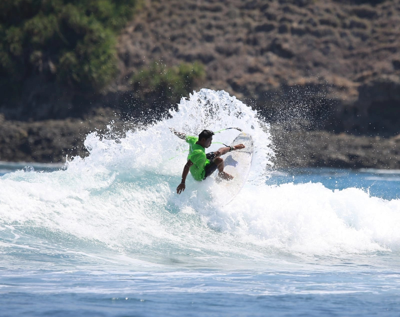 A male surfer carving on an ocean wave against a rugged coastal background.