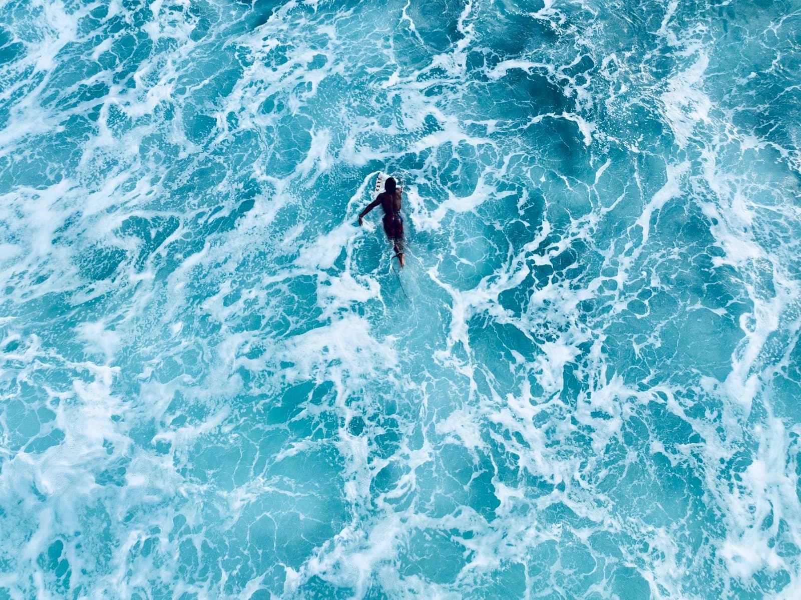 Aerial top-down view of a surfer paddling through foaming ocean water.