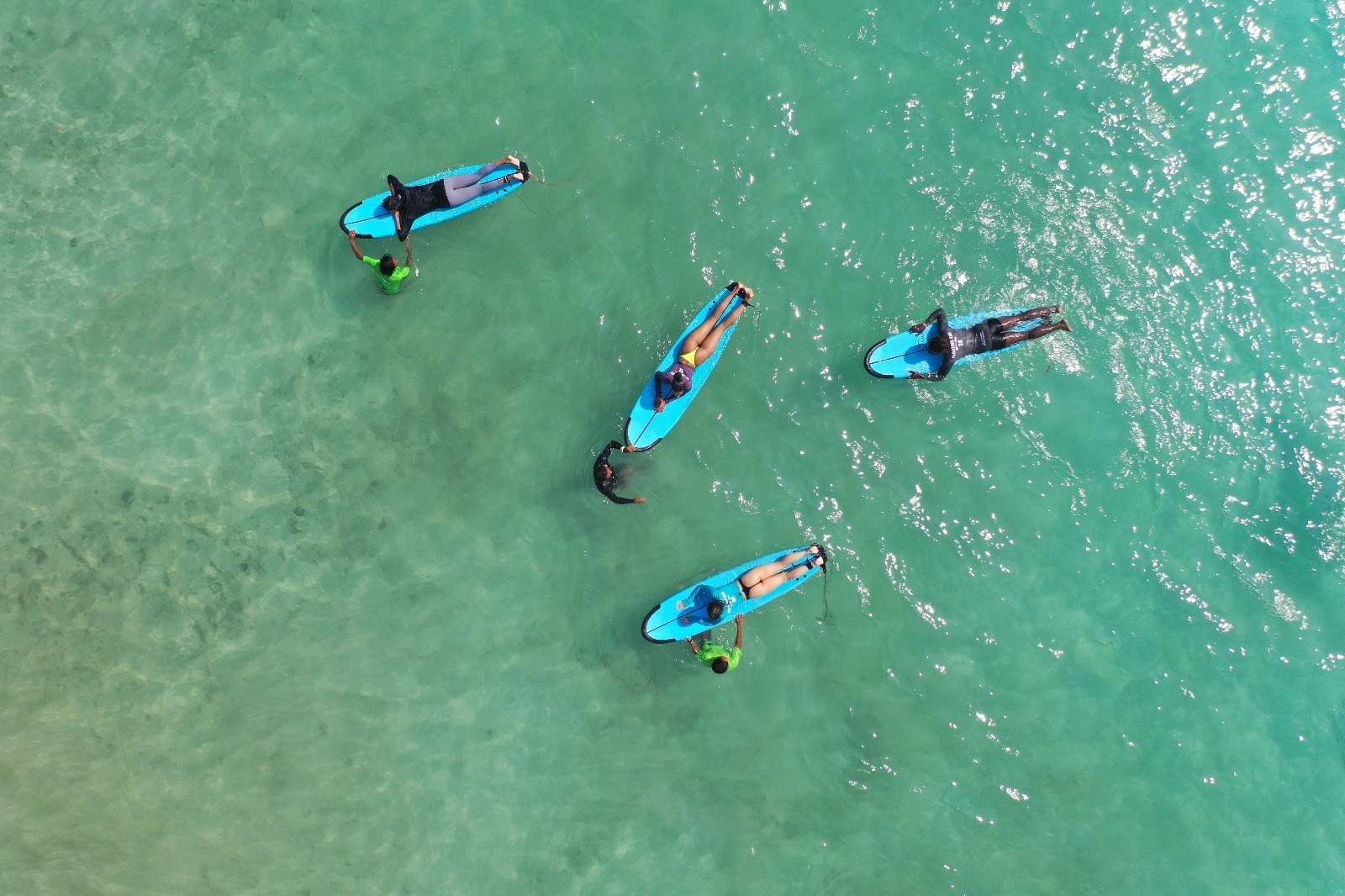 Aerial shot of surf instructors and students lying on blue foam longboards in clear turquoise sea water.