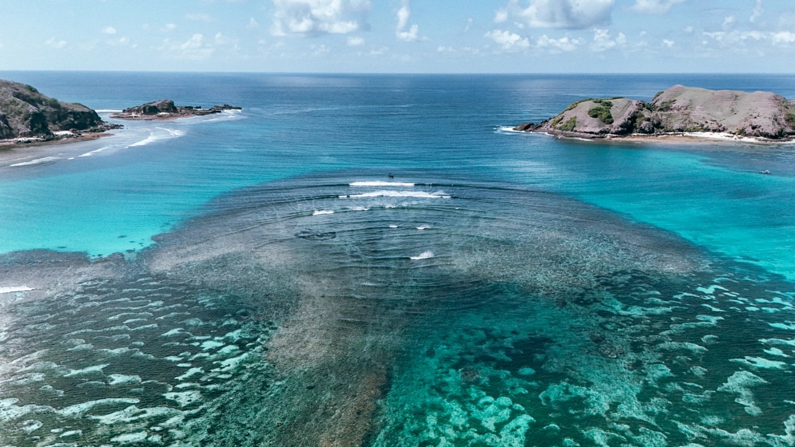 Wide aerial perspective of ocean swell breaking over a shallow reef between two coastal headlands.