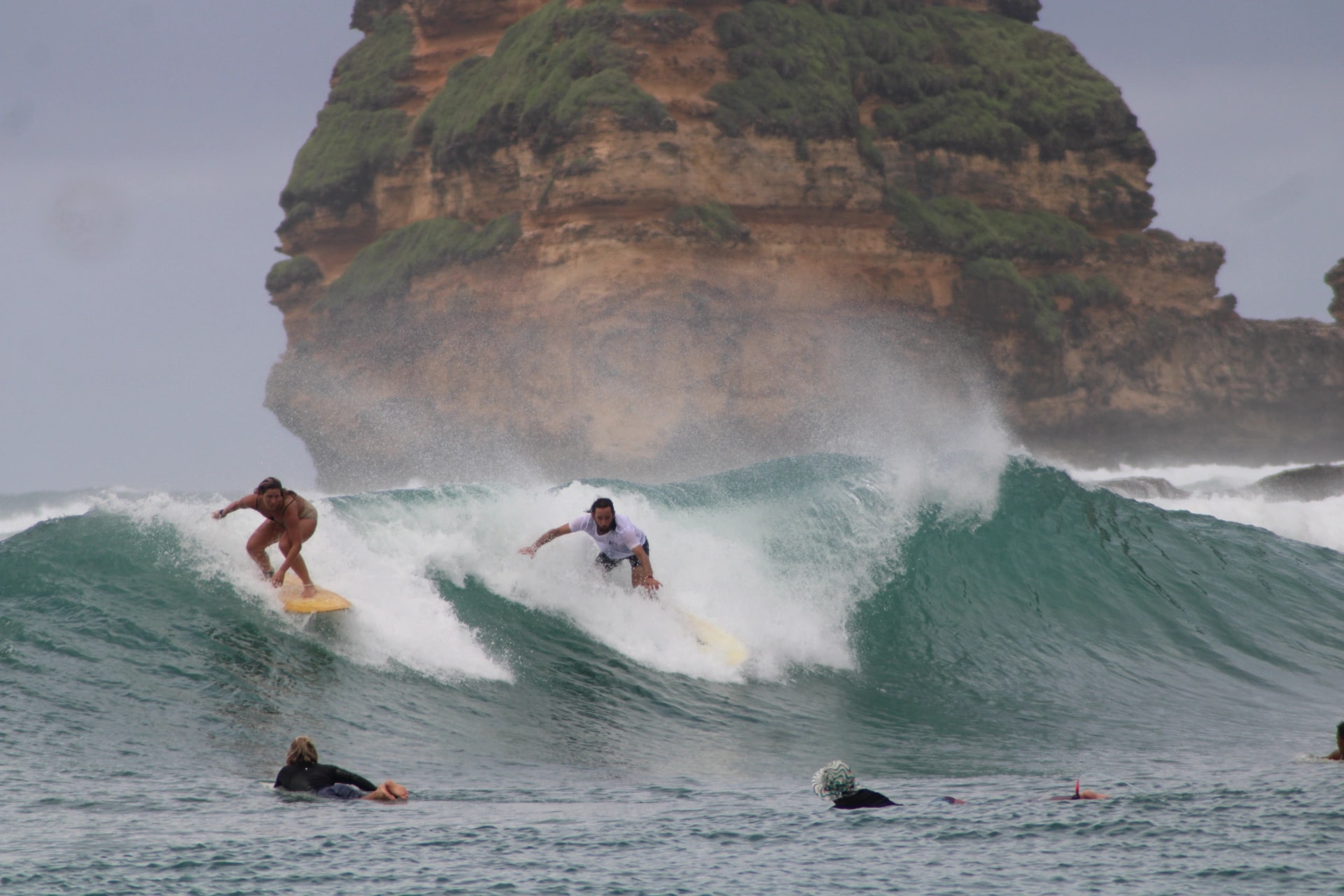 Surfers riding small waves near a large rocky sea stack covered in green foliage.