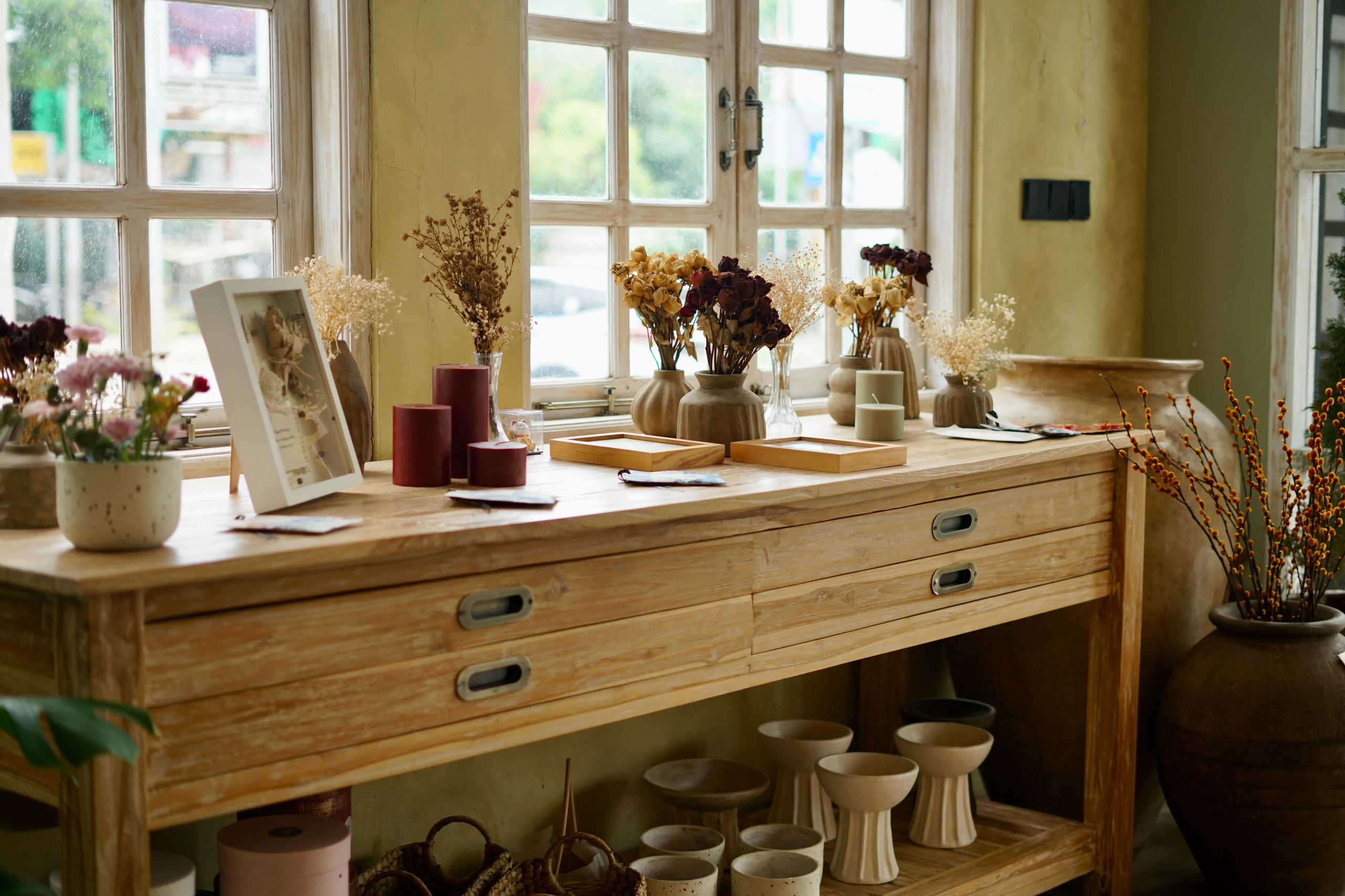 Wooden console table near a window decorated with candles, photo frames, dried flower vases, and ceramic pots.