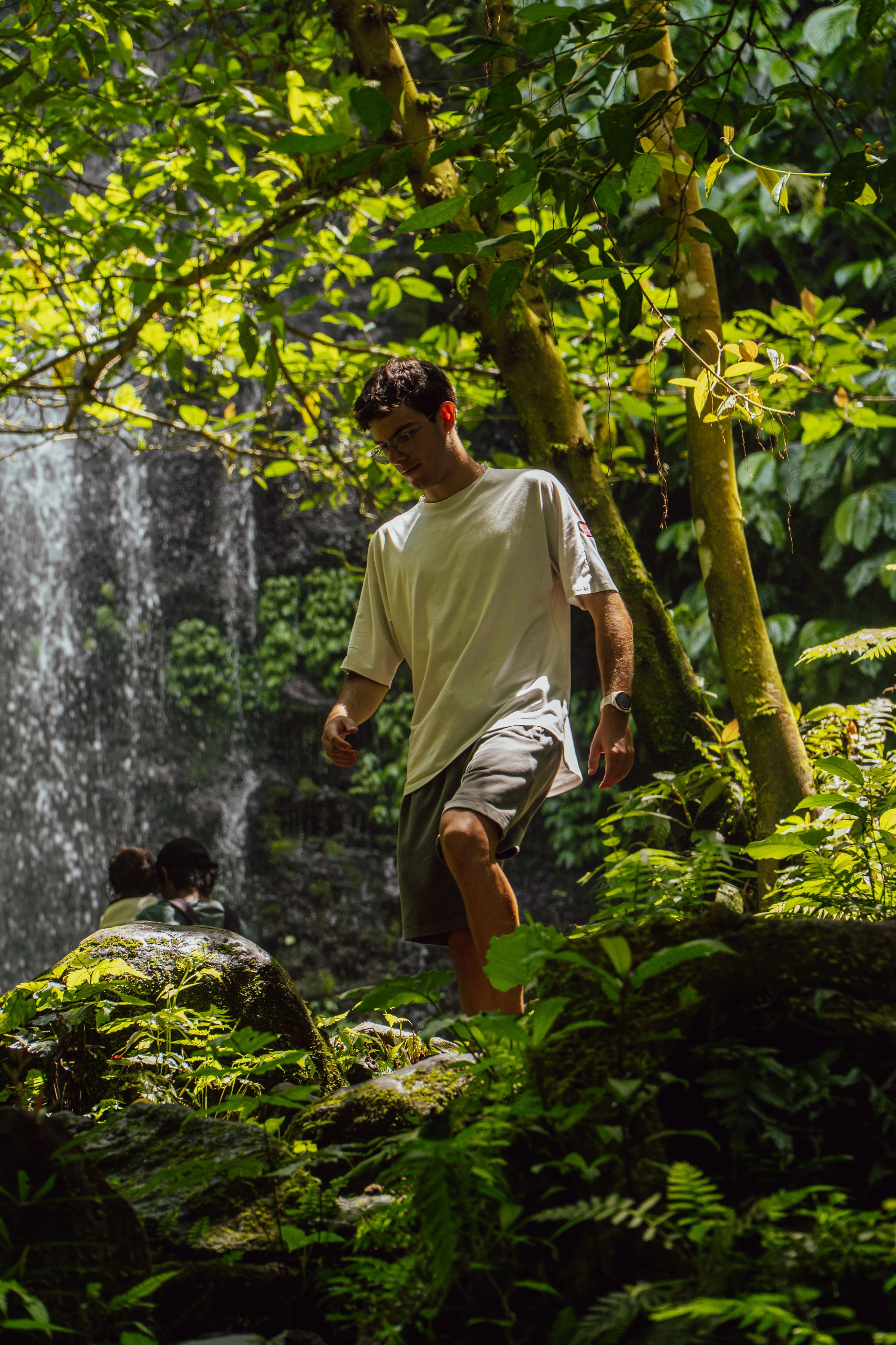 A young man wearing glasses and a white t-shirt steps down a rocky, forested path with a waterfall in the background.