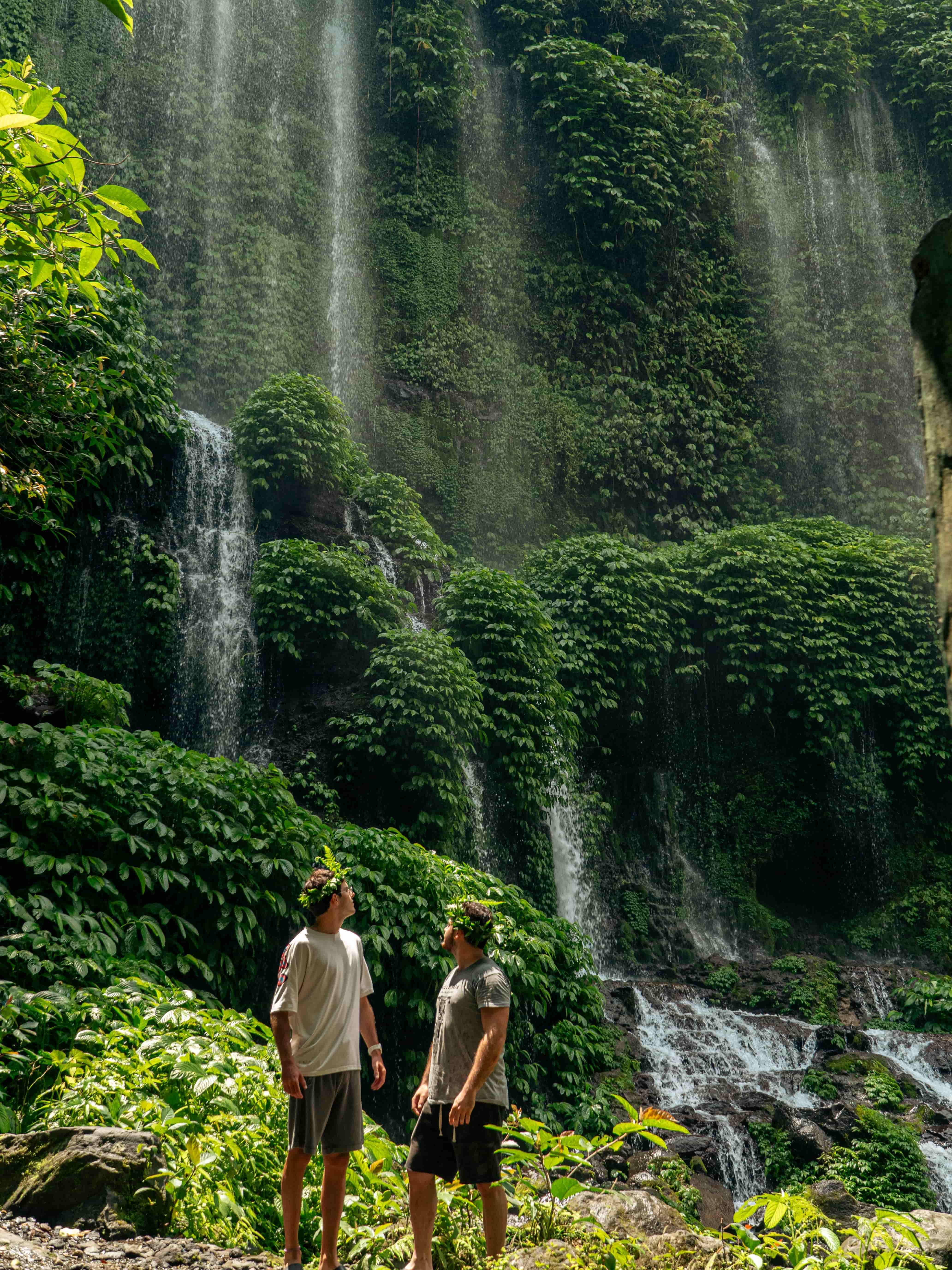 Two young men wearing leaf crowns stand and look up at a wide, curtain-like waterfall flowing down a lush, green cliff.