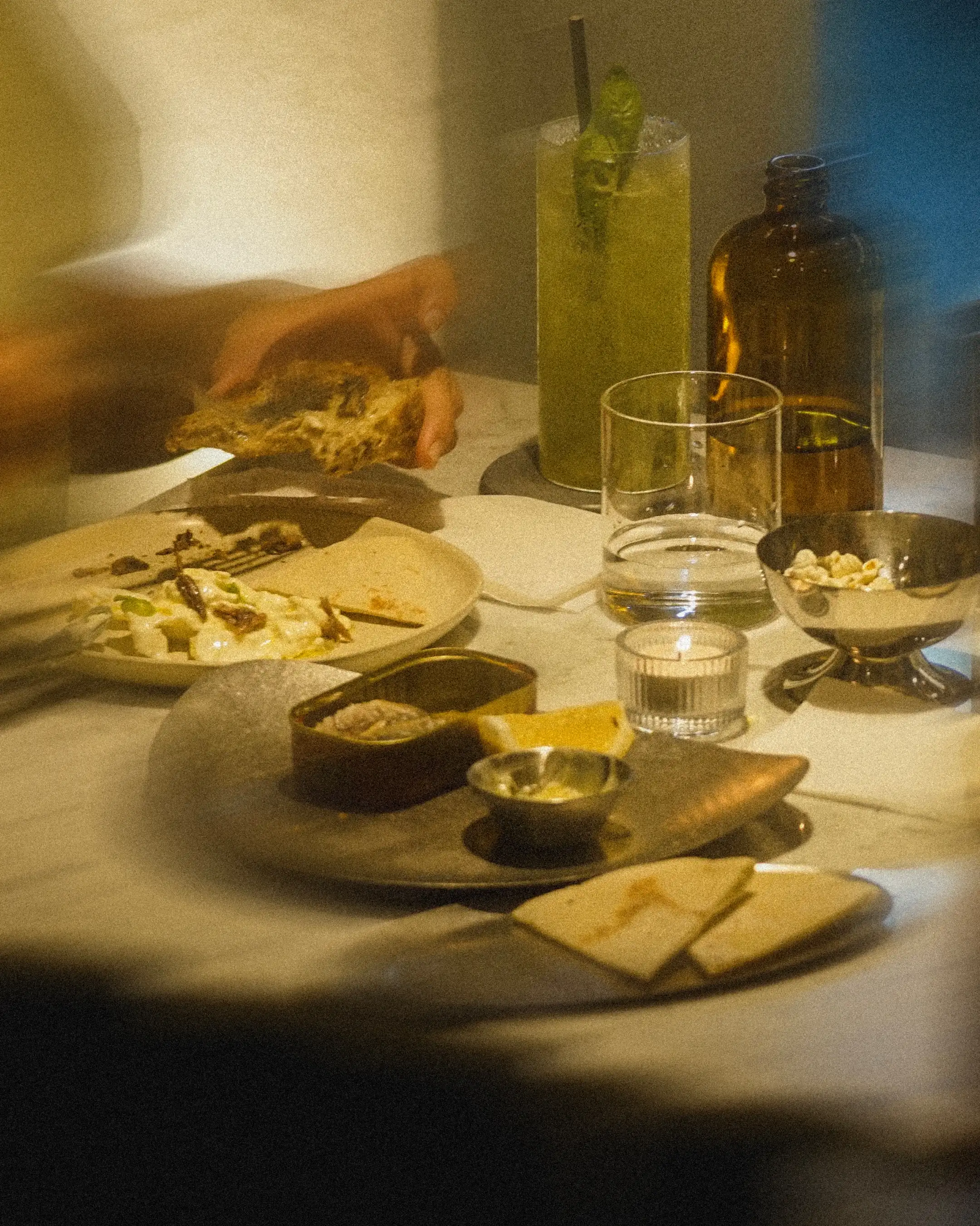 Close-up of a restaurant table featuring plated dishes, bread, sauces, drinks, and a refreshing green cocktail in a warmly lit setting.