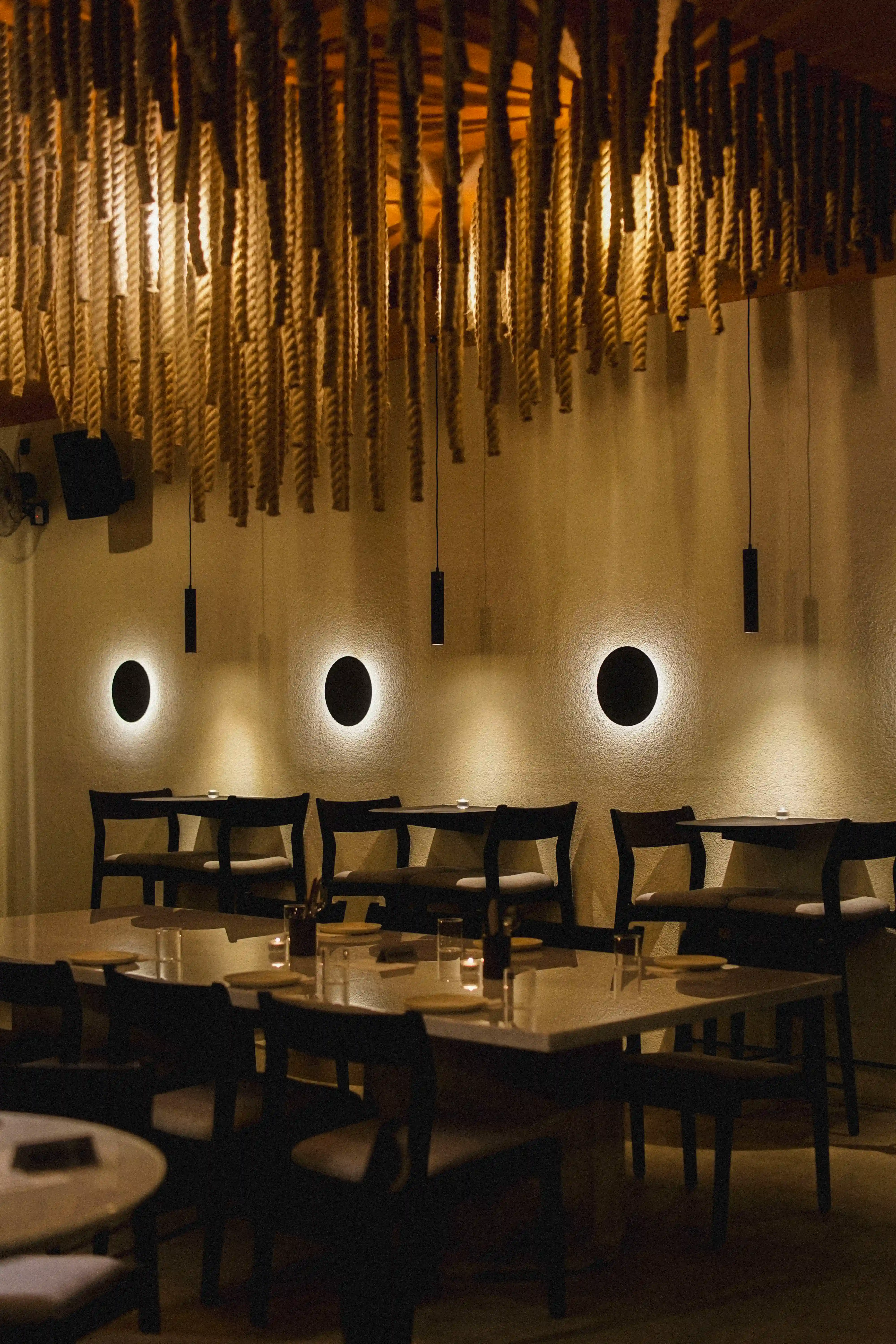 Elegant restaurant dining area with wooden tables, dark chairs, warm lighting, and decorative hanging rope lights.