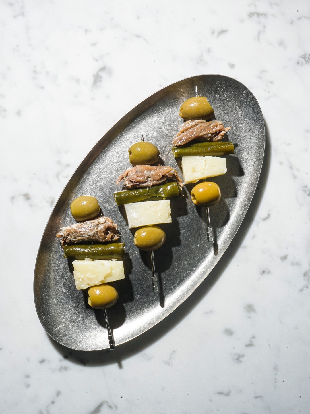 Three appetizer skewers with green olives, fish, and cheese served on an oval metal platter.