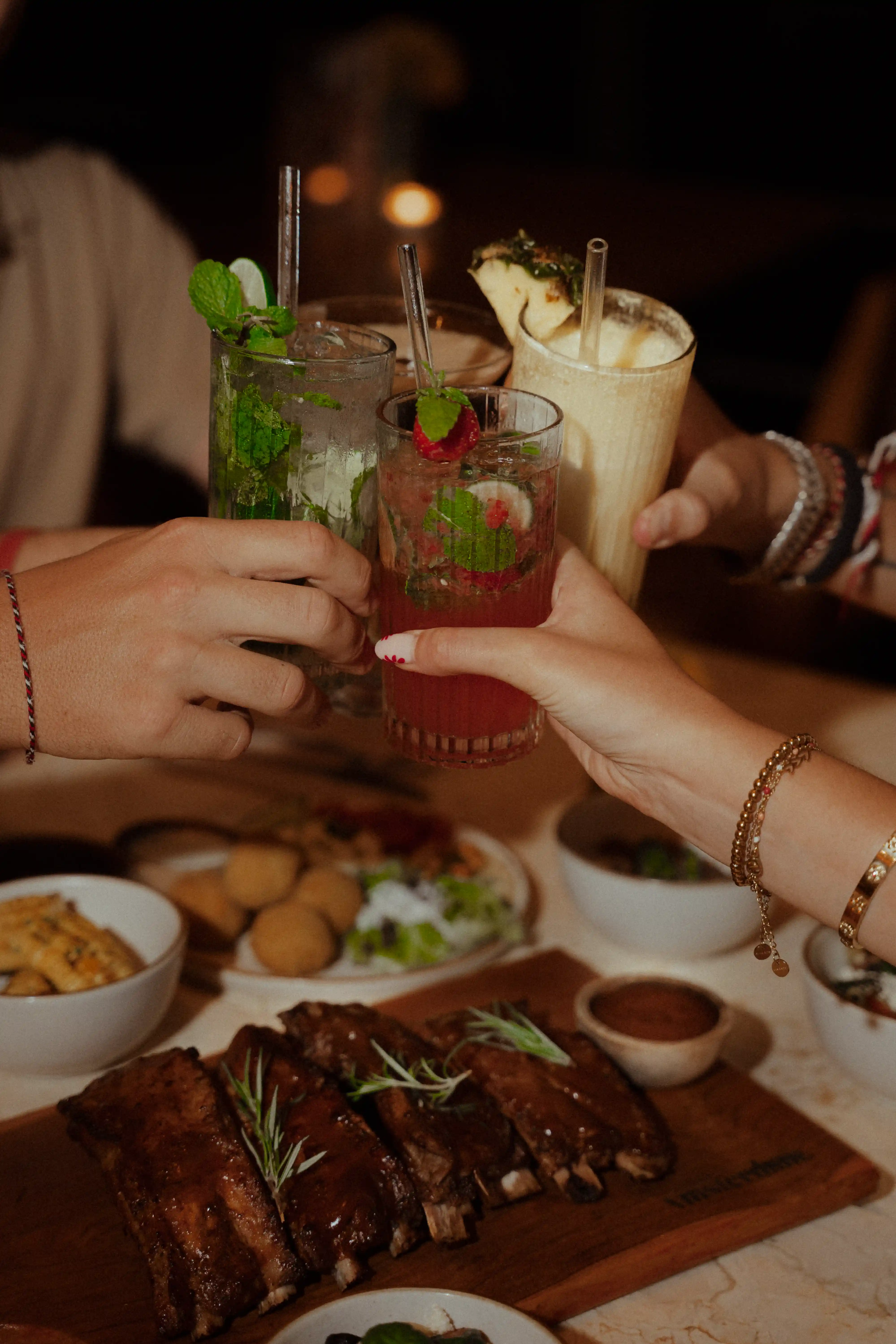 Close-up of people clinking cocktail glasses over a table served with BBQ ribs and side dishes.