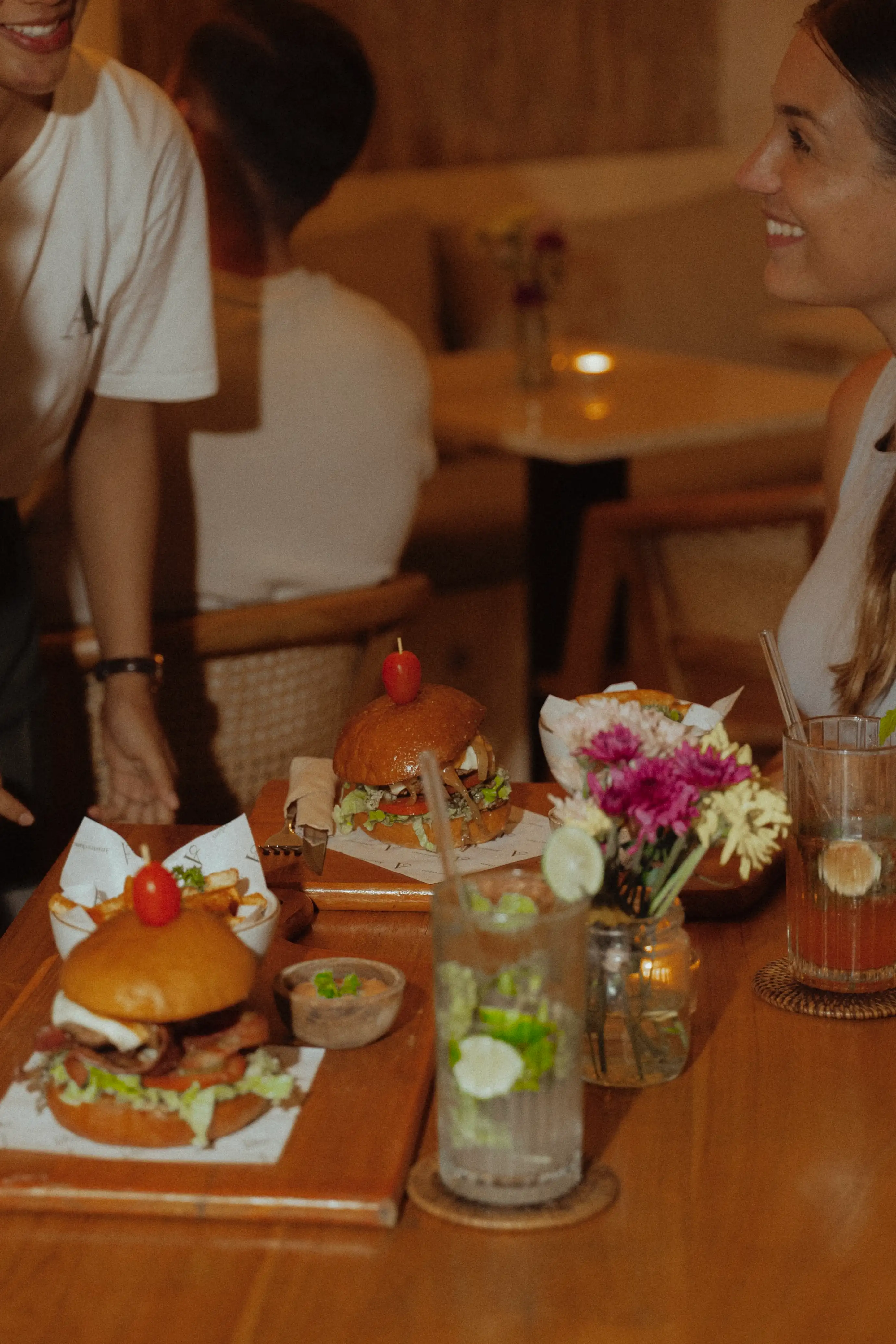 A woman smiling at a server while burgers, fries, and cold drinks are served on a wooden table.