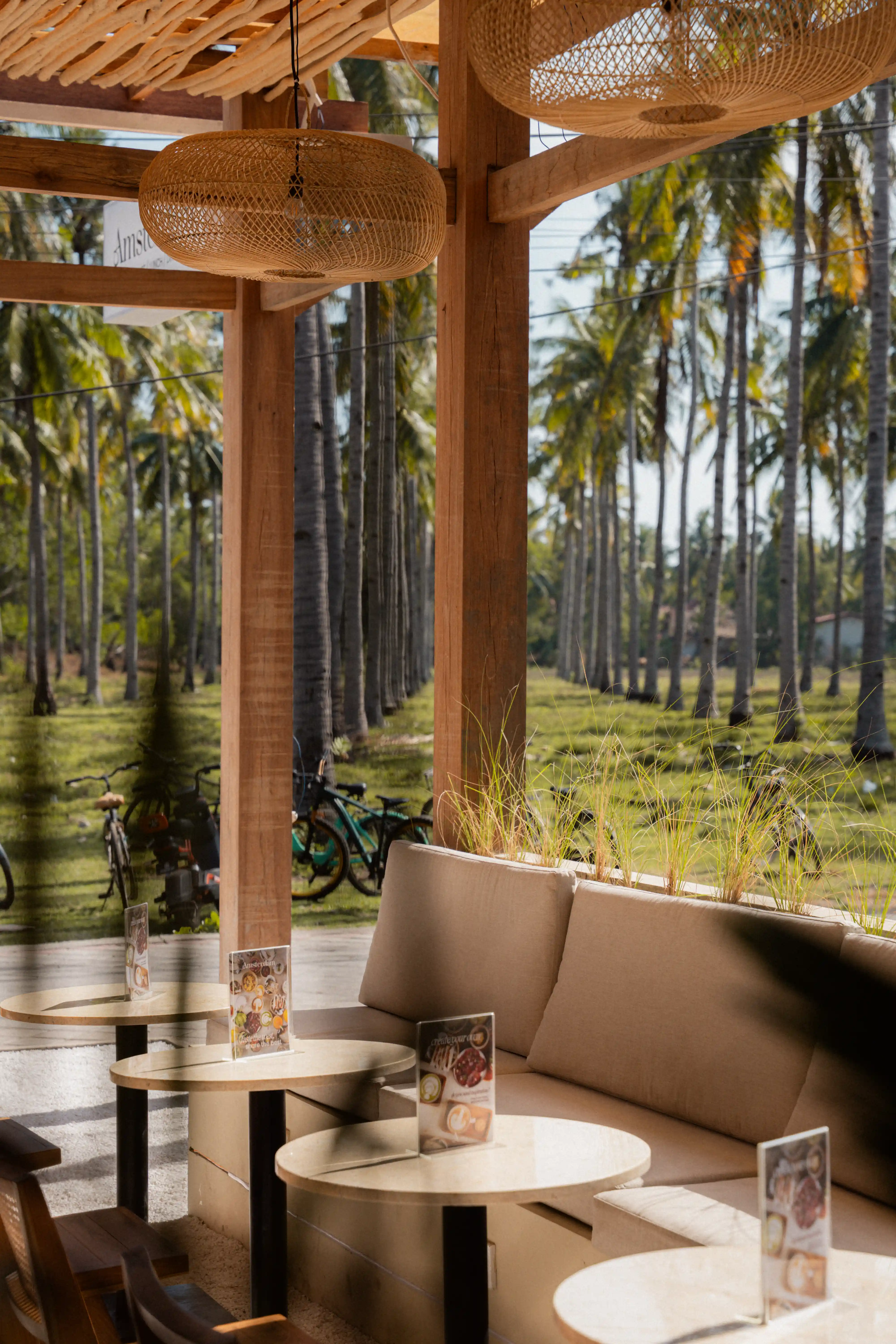 Open-air restaurant seating area with beige couches and round tables overlooking a tropical palm field.