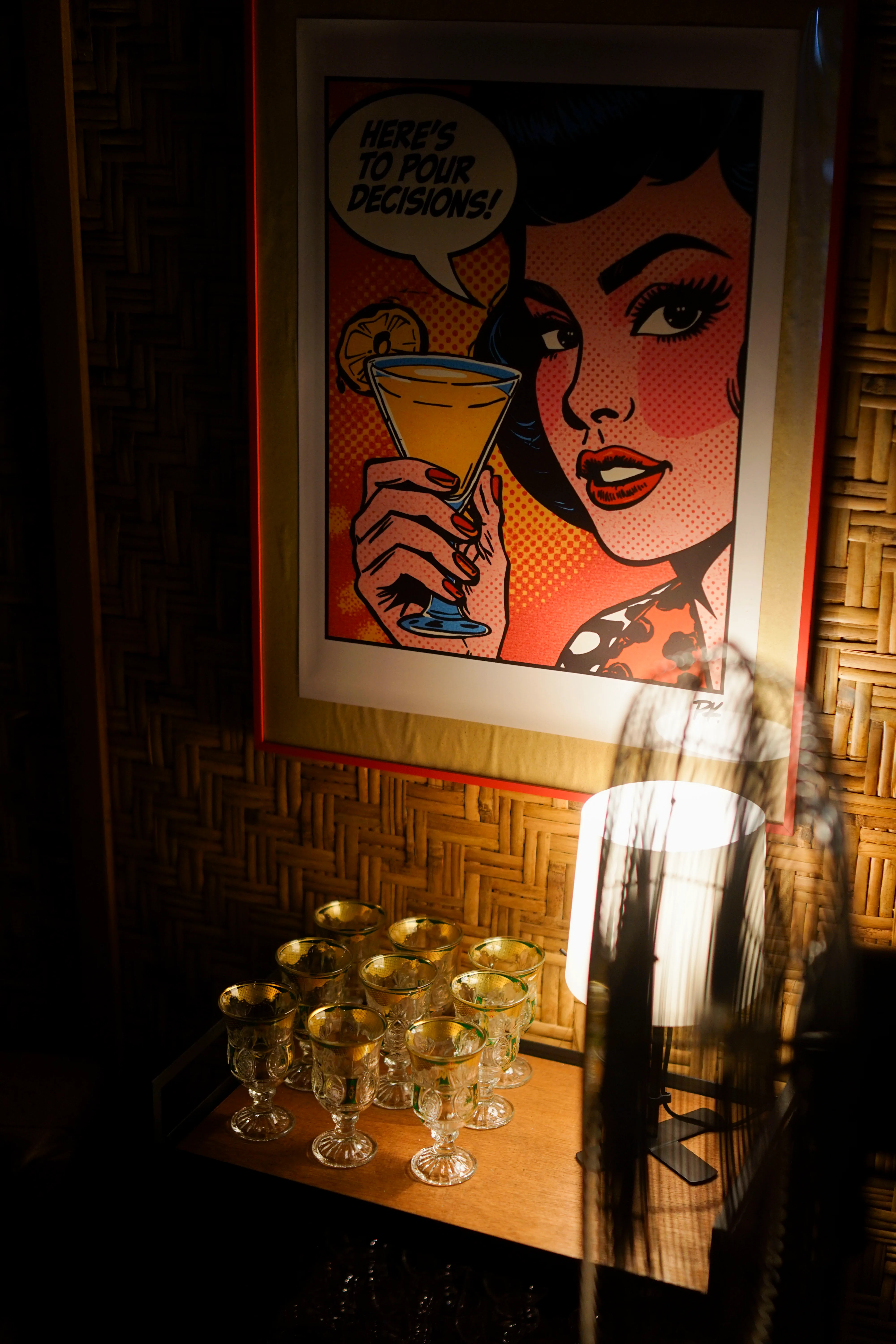 Framed pop-art poster reading "Here's to pour decisions!" above vintage glasses and a lamp on a wooden shelf.