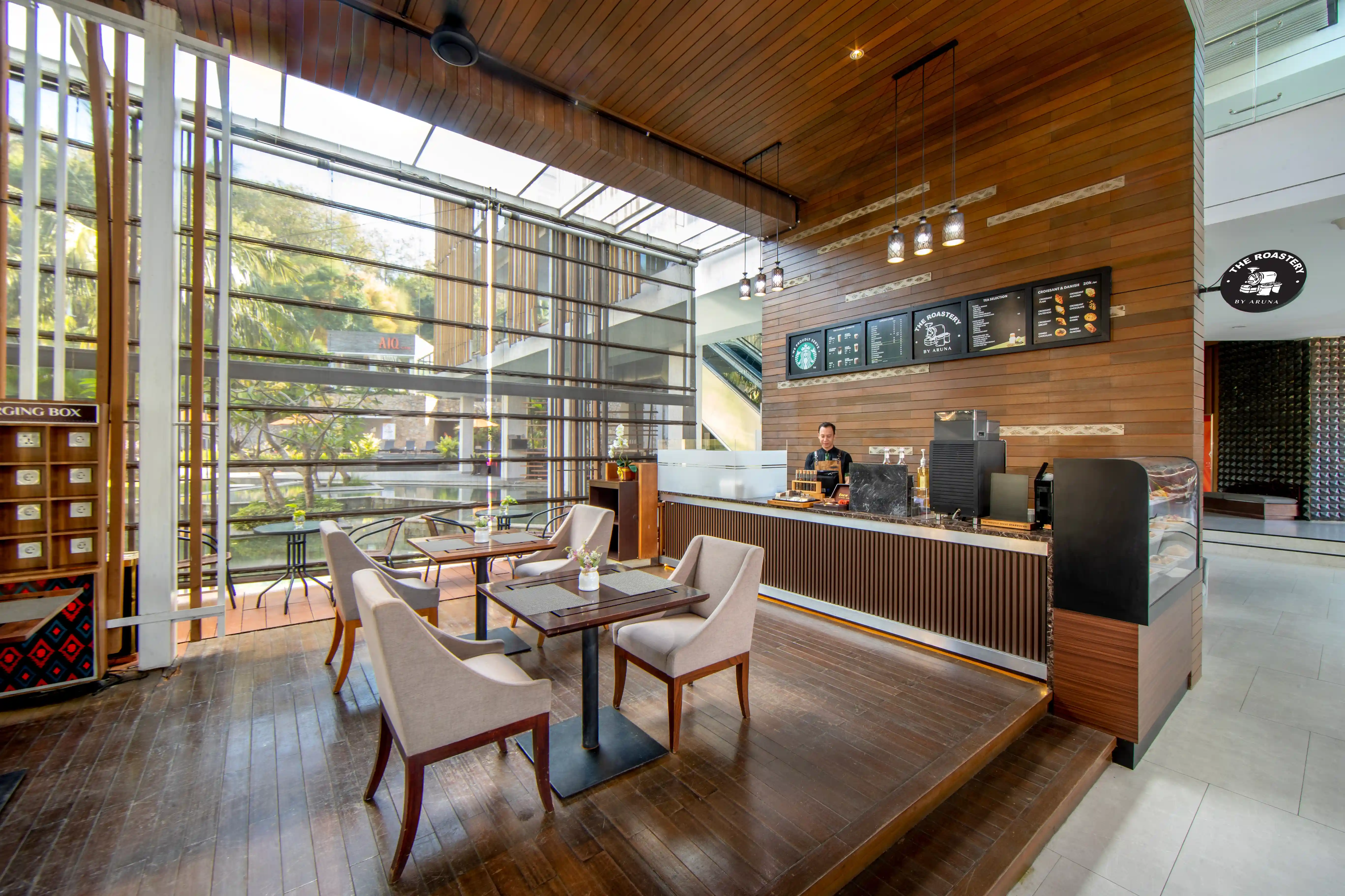 Modern café, The Roastery, with a wooden counter, floor-to-ceiling windows, and dining tables.