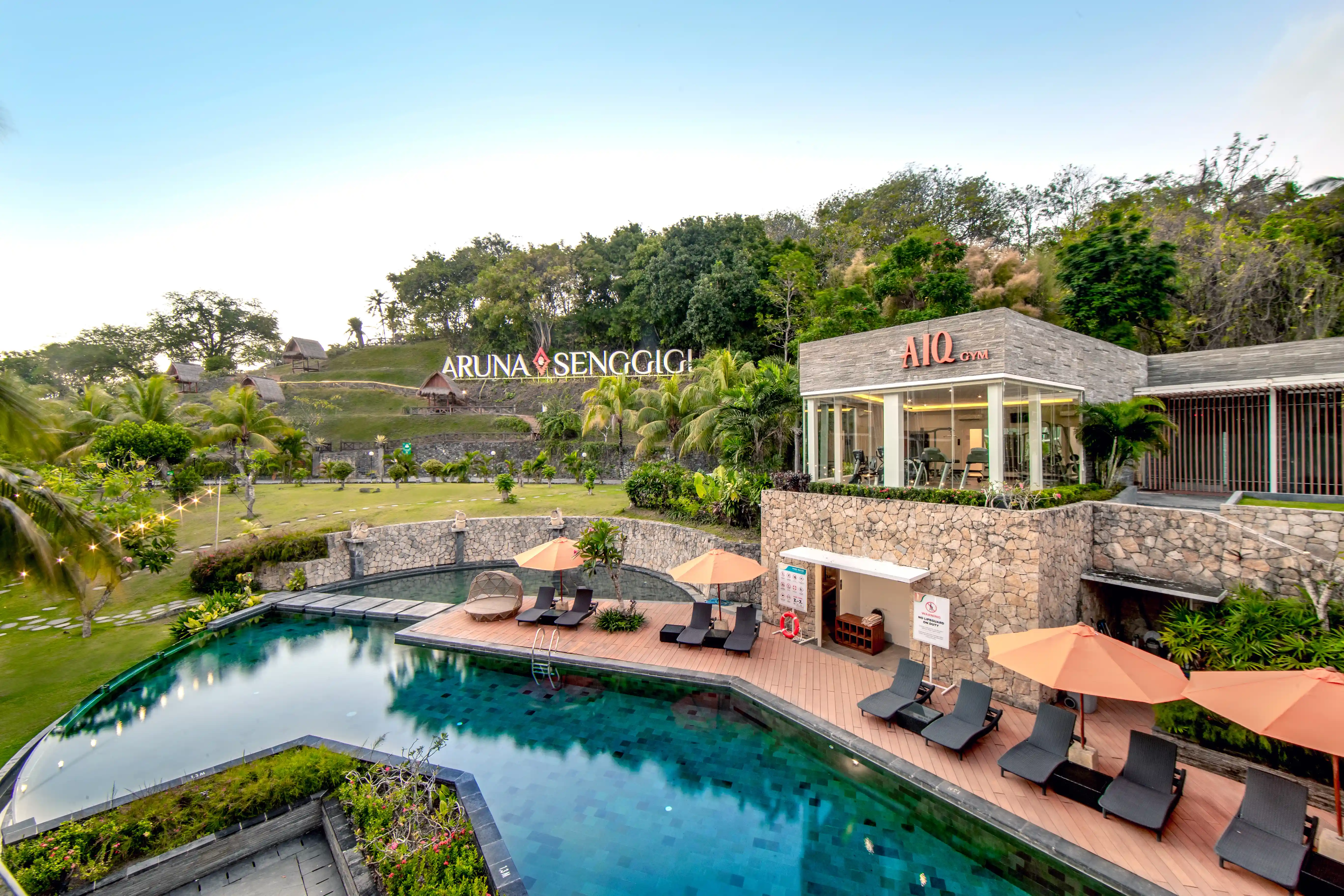 Outdoor hotel pool area with sun loungers, orange umbrellas, and the gym building in the background.