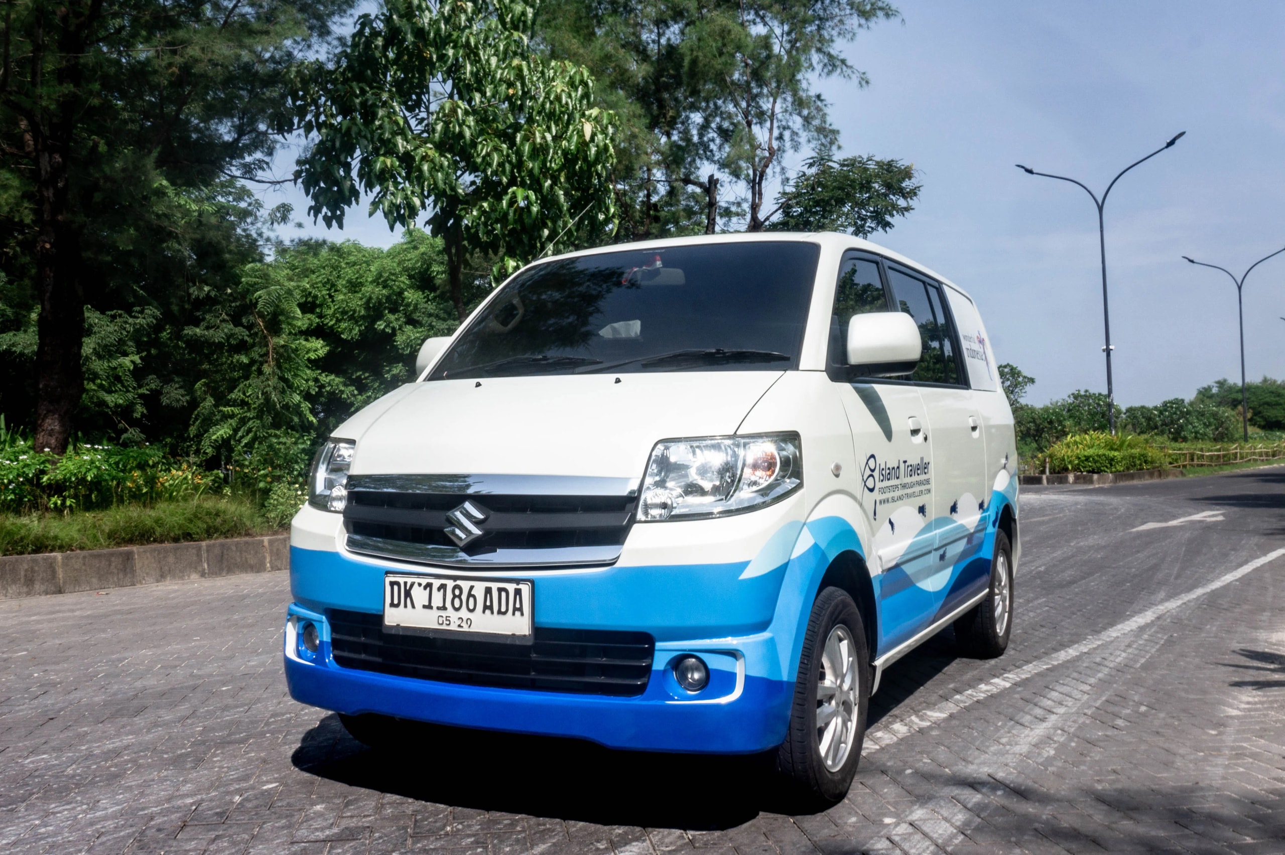 Front-three-quarter view of a white Suzuki passenger van with blue ocean-wave decals parked on a paved driveway.