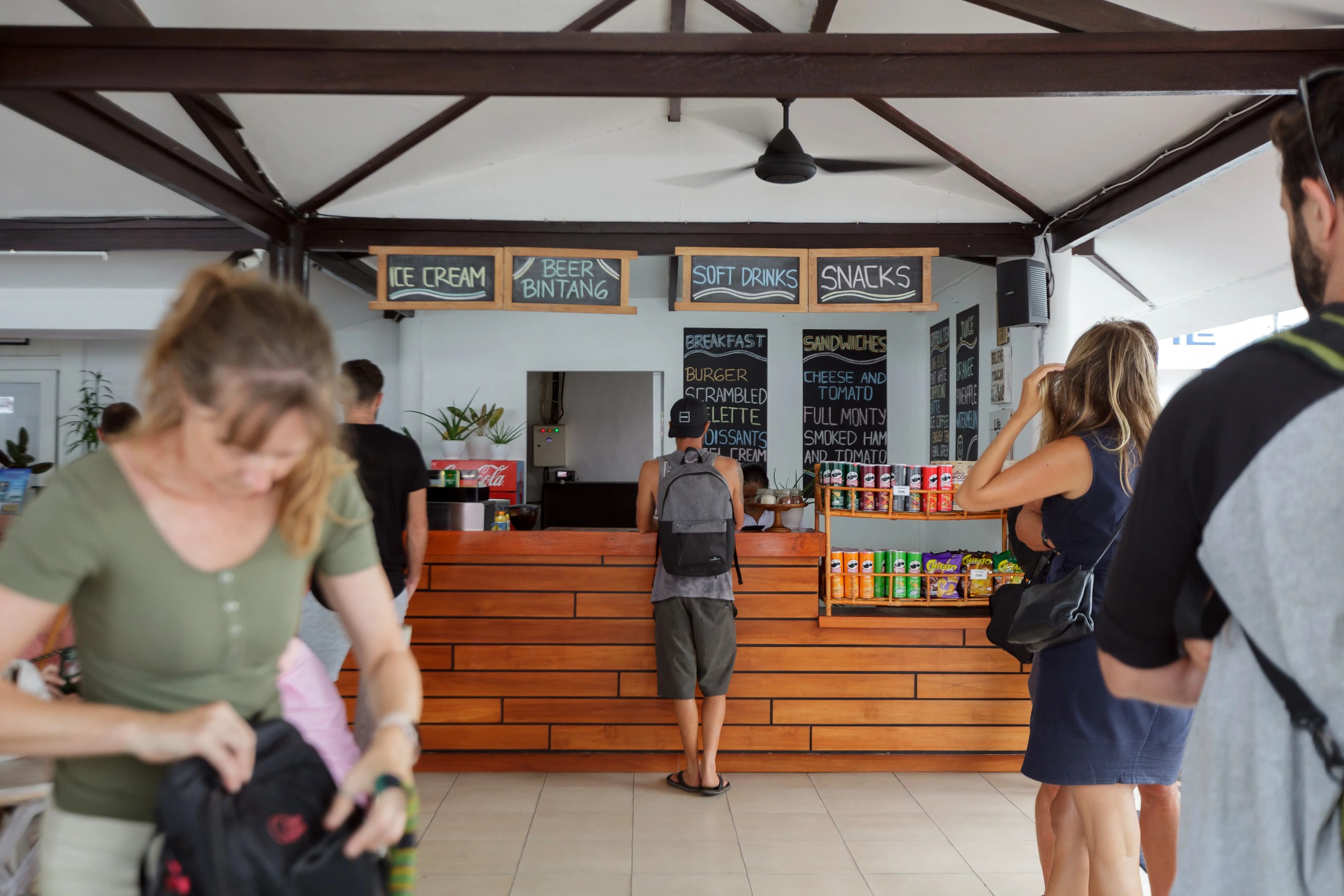 Open-air harbor snack bar counter with chalkboard menus, snacks on display, and several tourists ordering or waiting nearby.