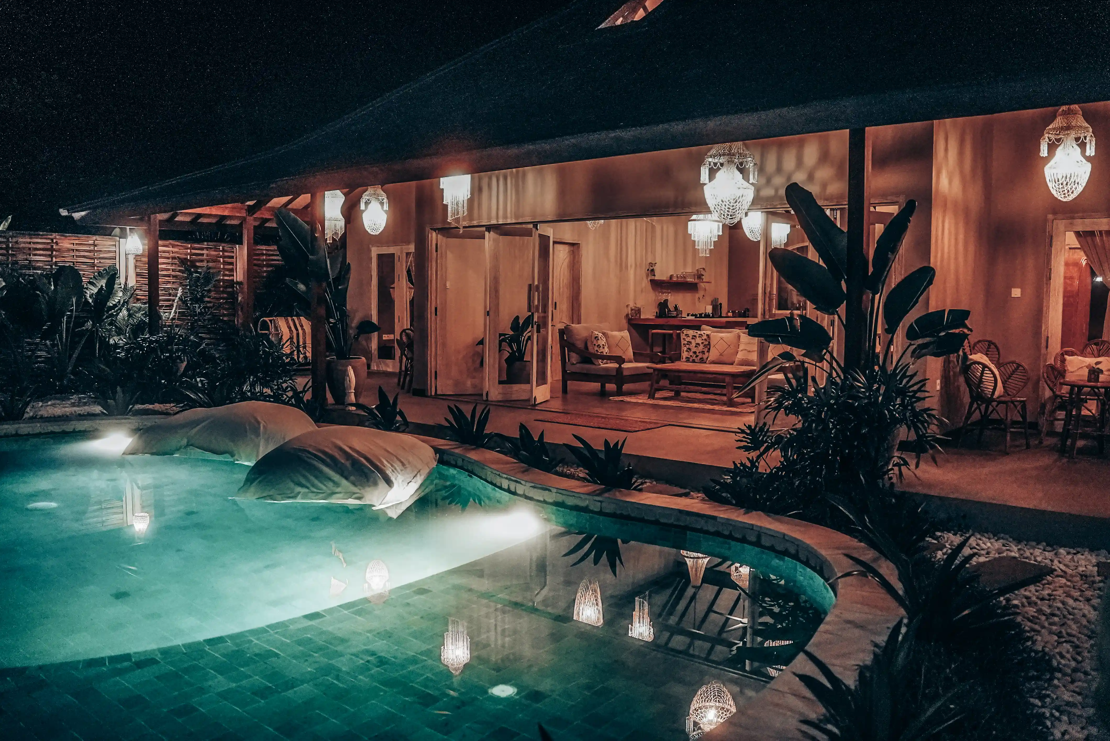 Vue nocturne de la villa éclairée avec une piscine extérieure bordée de plantes tropicales.