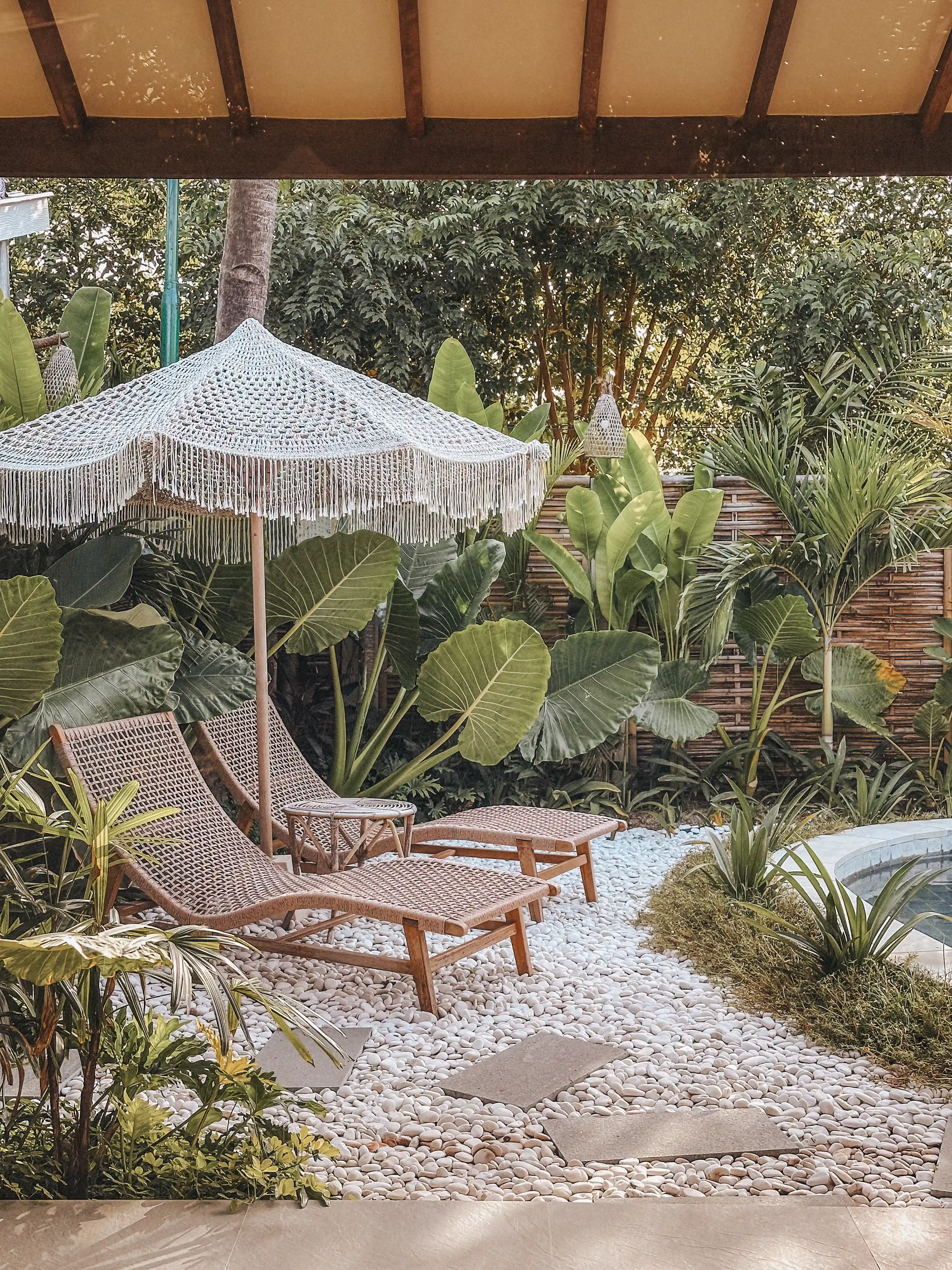 Coin détente extérieur au bord de la piscine avec transats en rotin et parasol à franges sur sol de galets blancs.