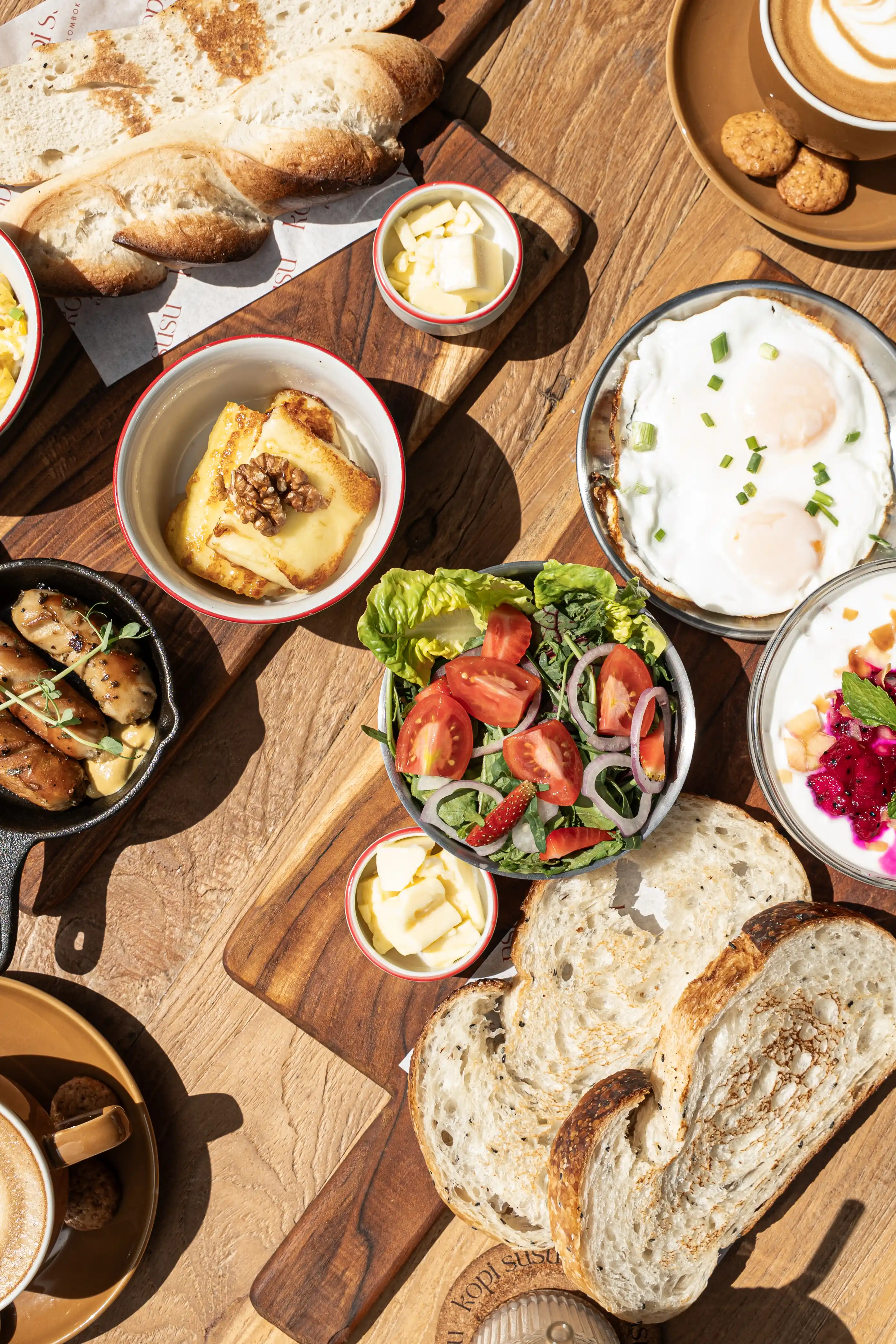 Overhead view of a breakfast spread with bread, eggs, salad, yogurt, butter, and coffee on a wooden table.