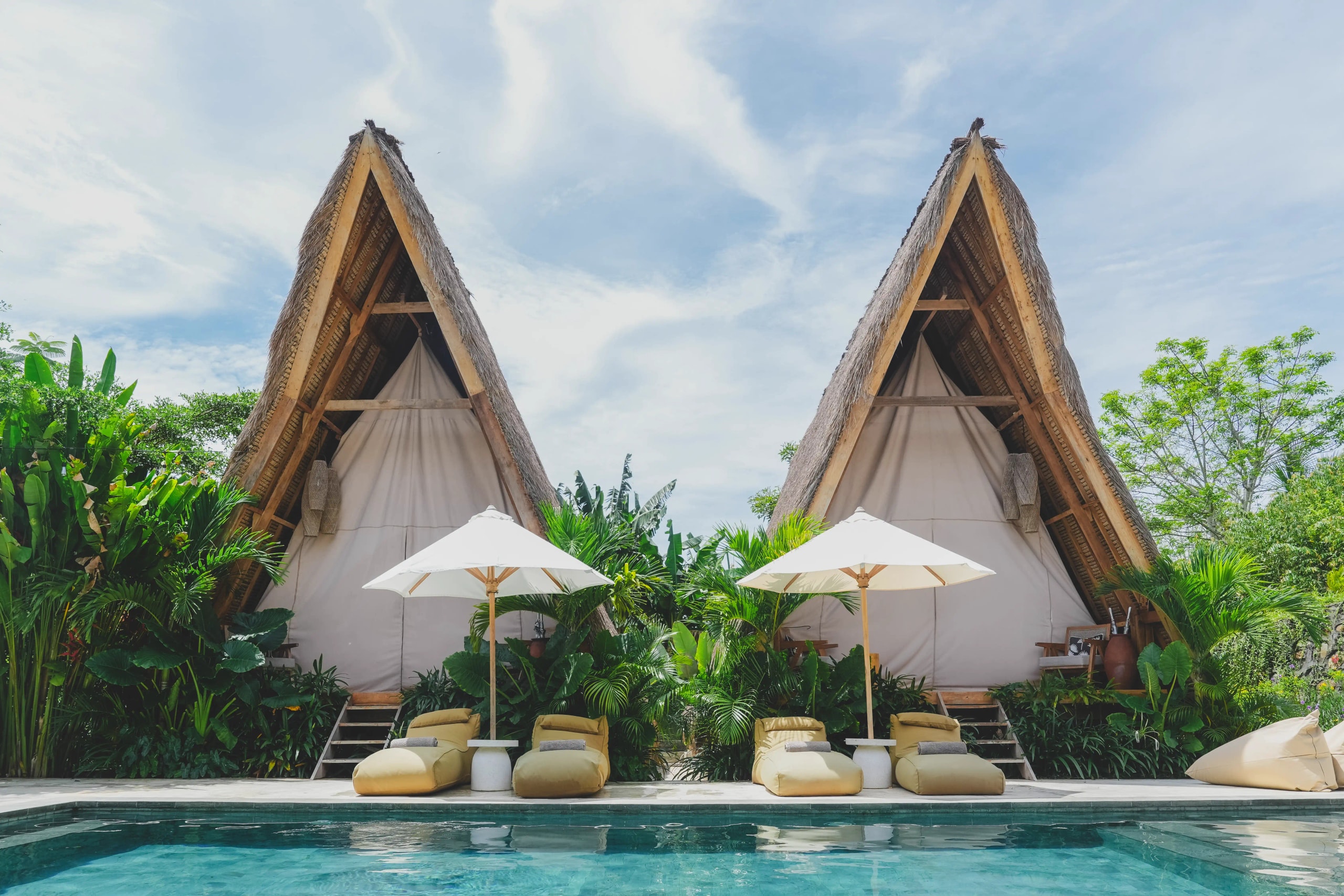 Two thatched-roof A-frame glamping tents behind a swimming pool lined with cushioned sun loungers and white umbrellas.