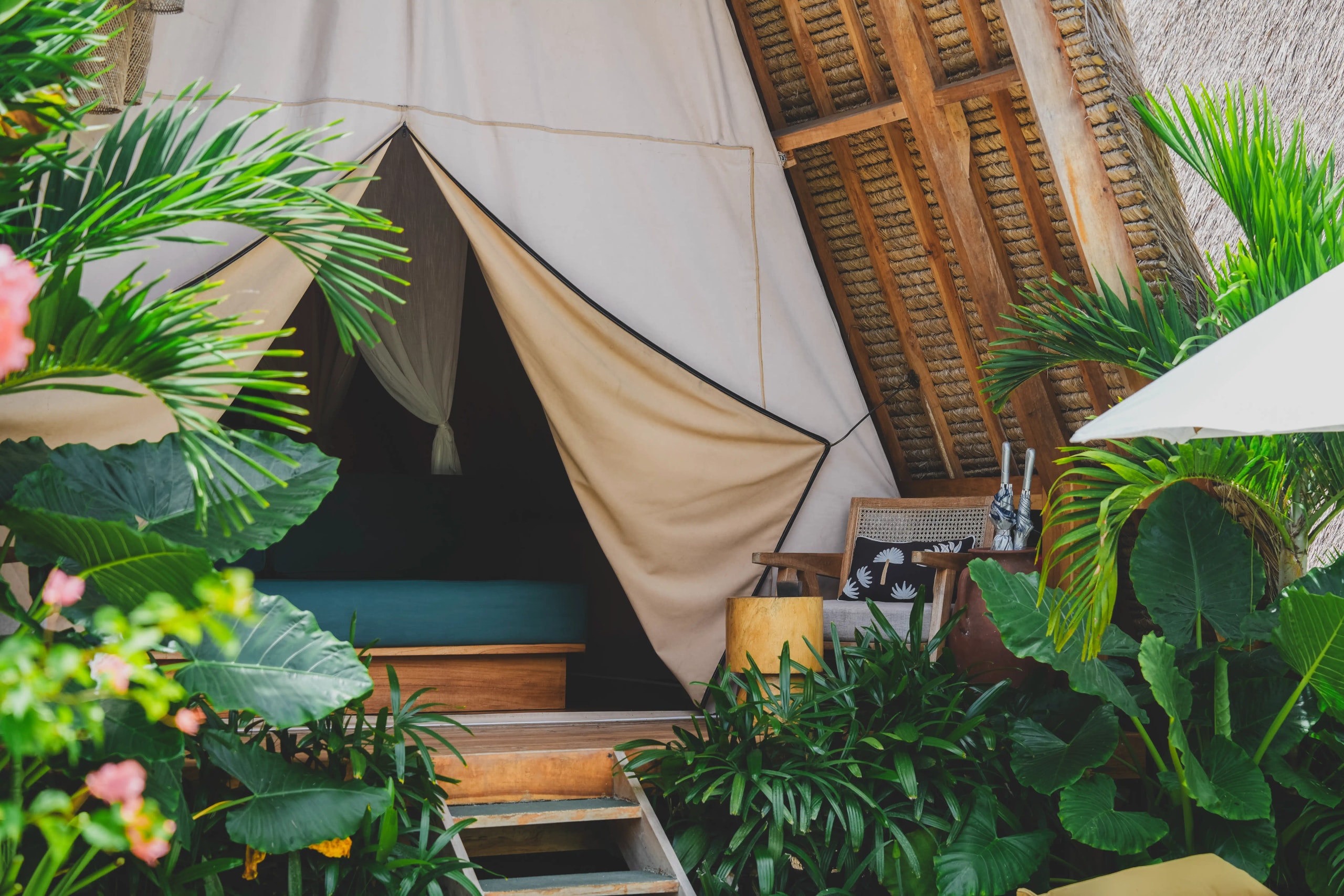 Entrance to a luxury glamping tent with a wooden deck, armchair, and lush tropical plants.