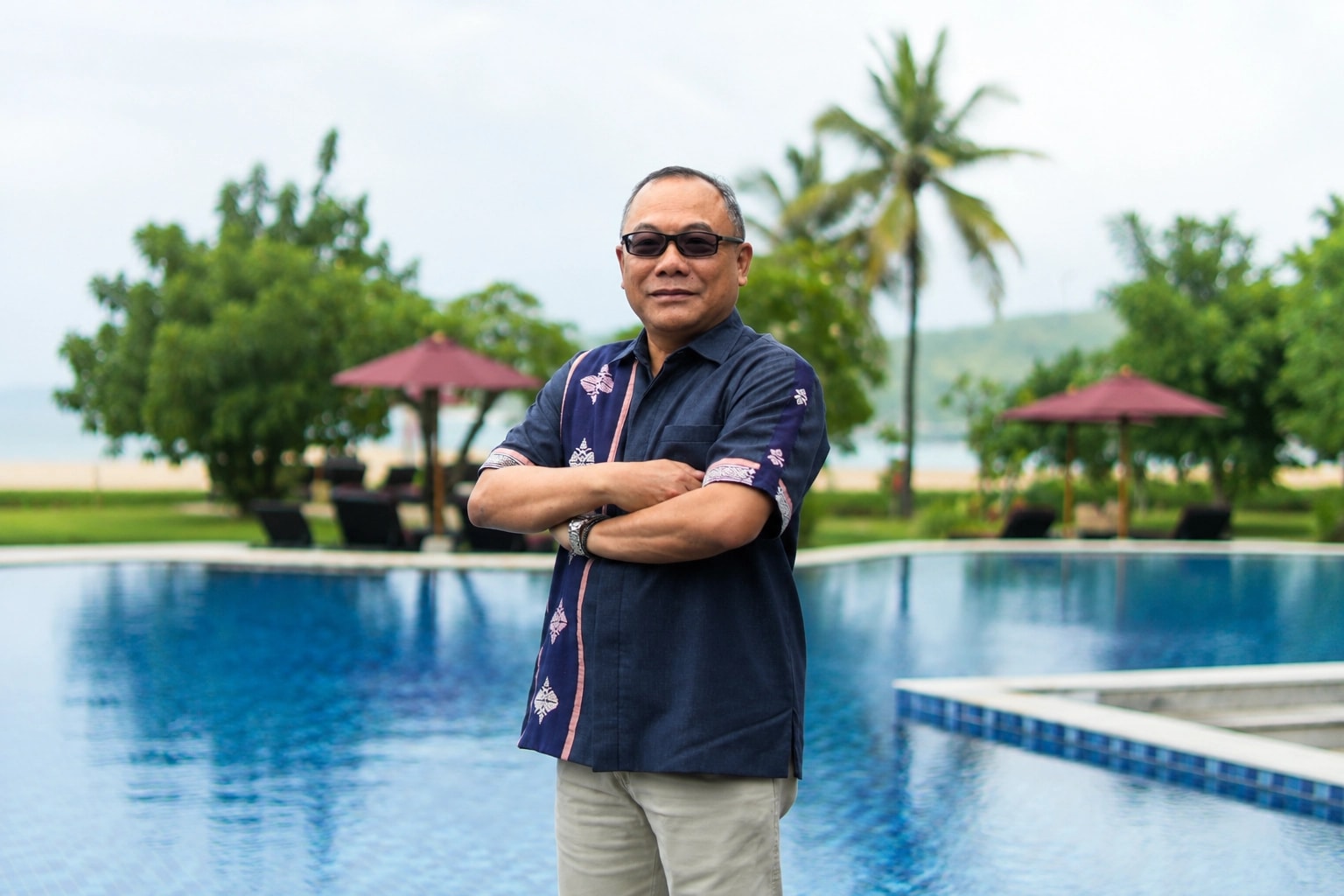 Man in a patterned navy shirt with arms crossed standing by an outdoor swimming pool at a beach resort.