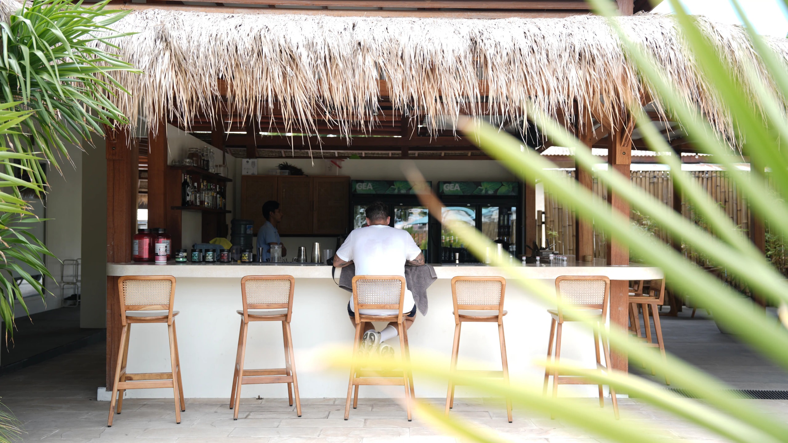 Open-air tiki bar featuring a thatched roof, wooden high-top stools, and a customer sitting at the counter.