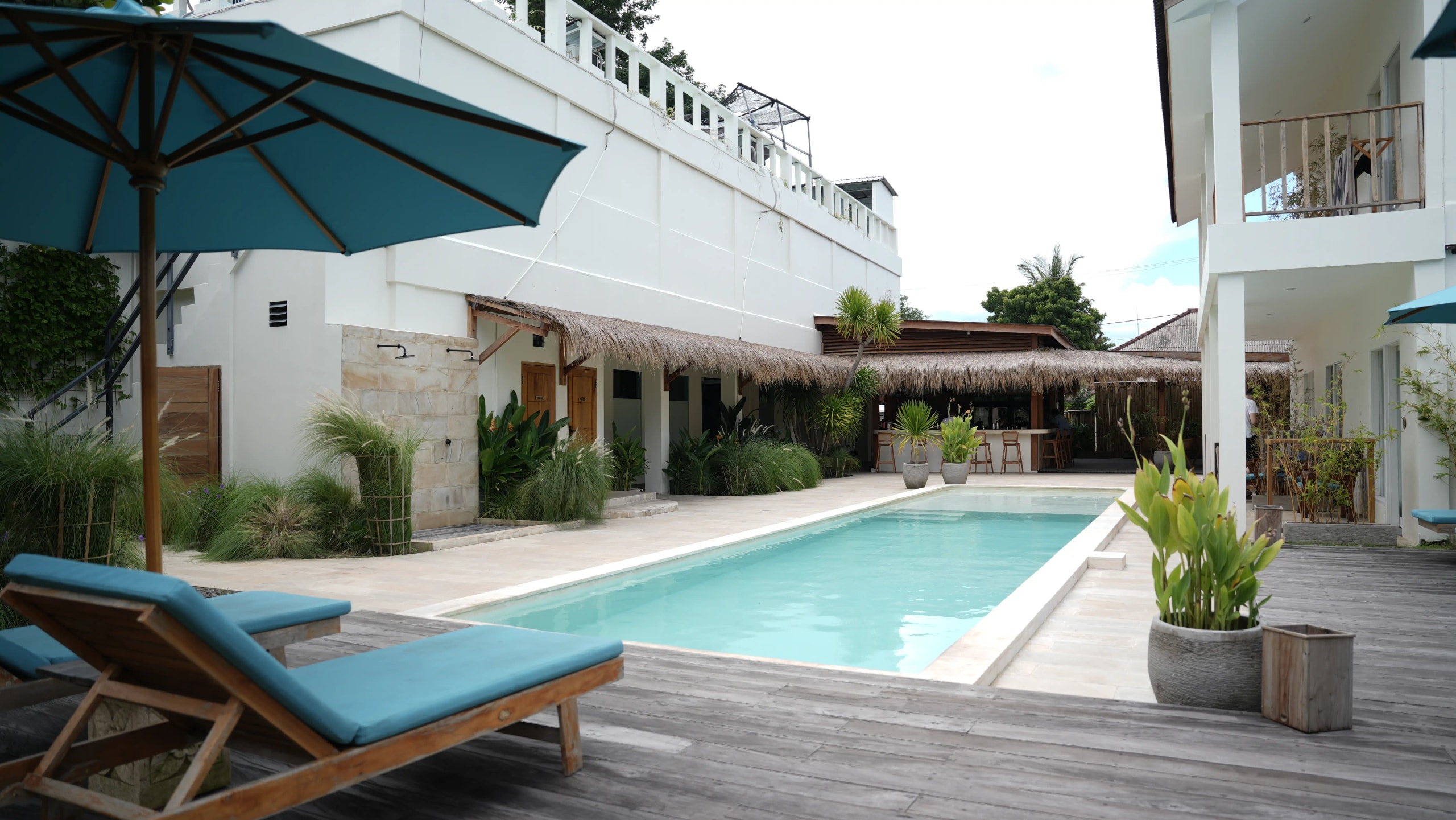Courtyard area featuring a long outdoor swimming pool flanked by wooden sun loungers, umbrella shading, and modern resort buildings.