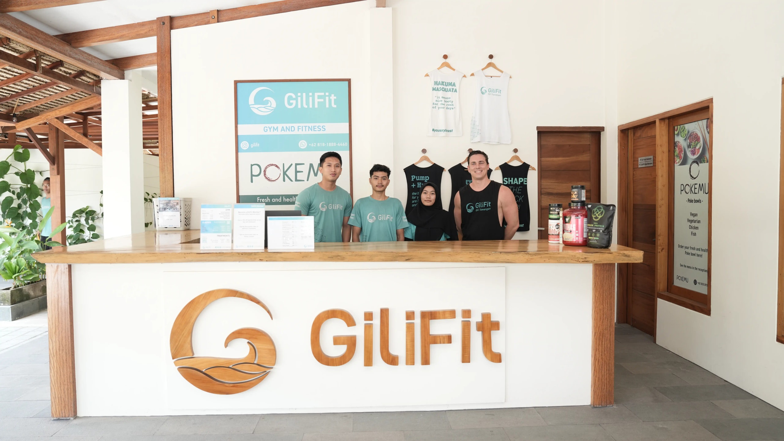 Front reception counter of GiliFit with wooden branding, where four staff members in branded shirts stand smiling behind the desk.