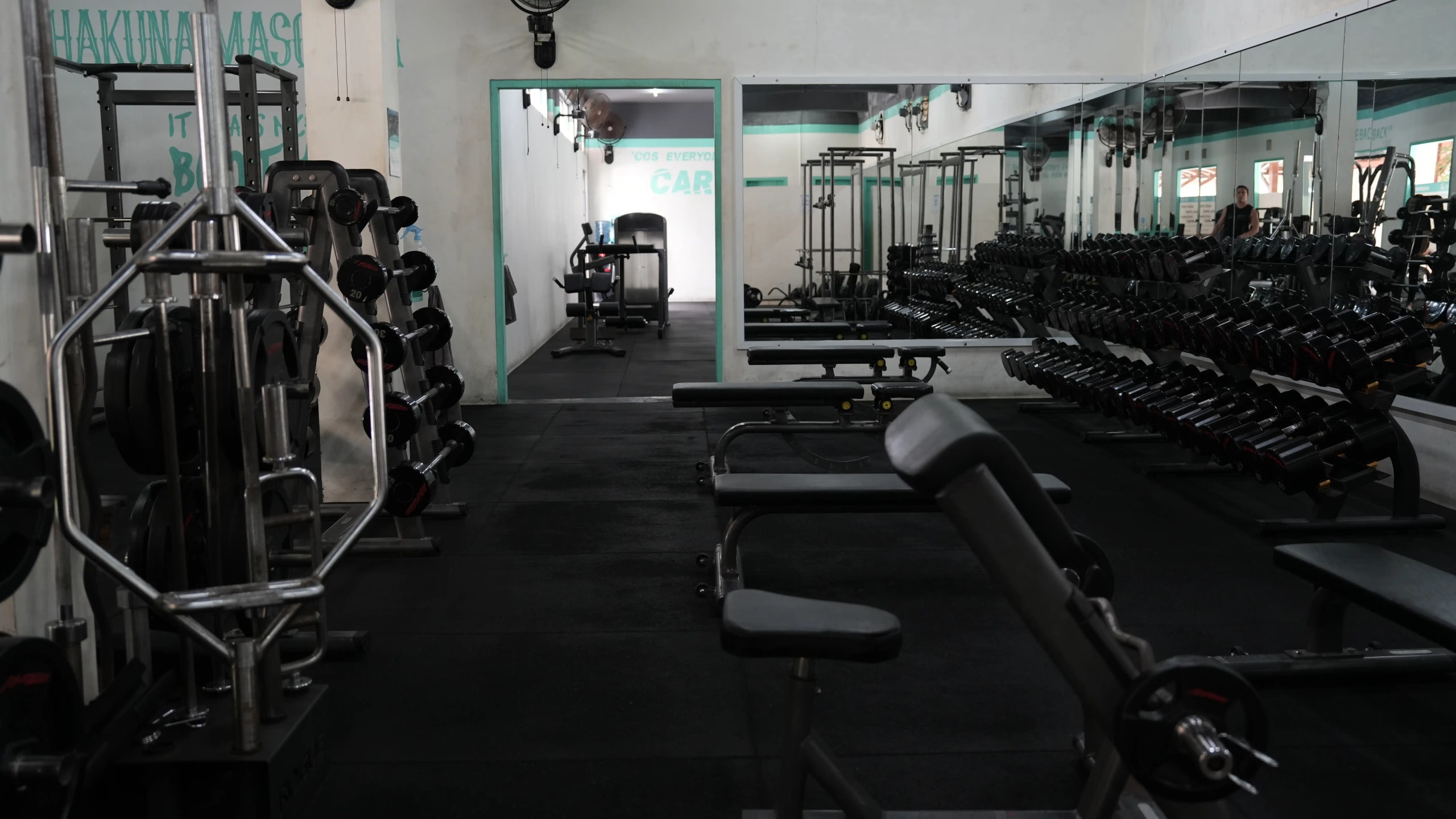 Interior view of an indoor weight room equipped with dumbbell racks, weight benches, barbell squat racks, and wall mirrors.