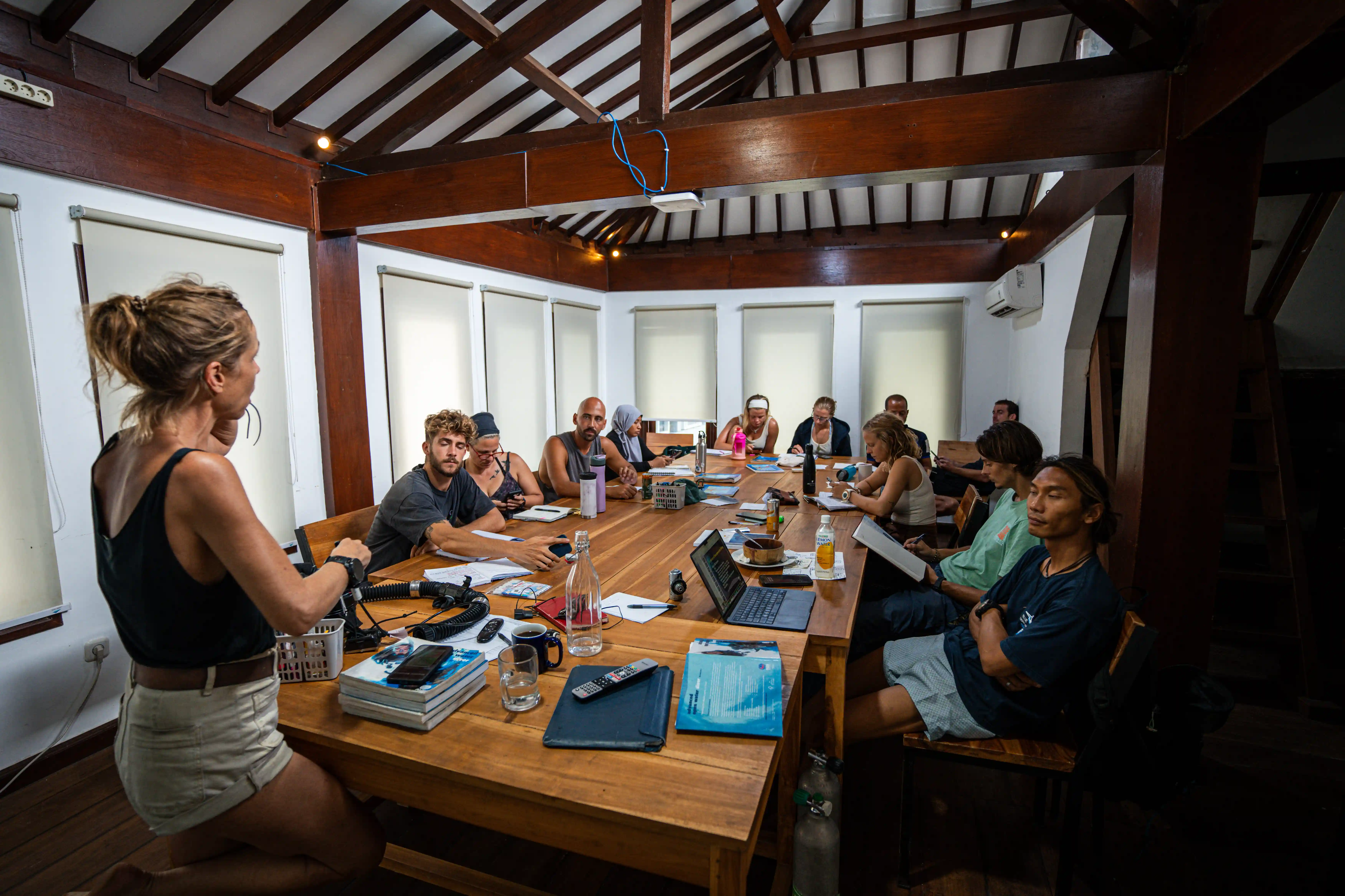 Scuba diving instructor leading a classroom session with students seated around a long wooden table.