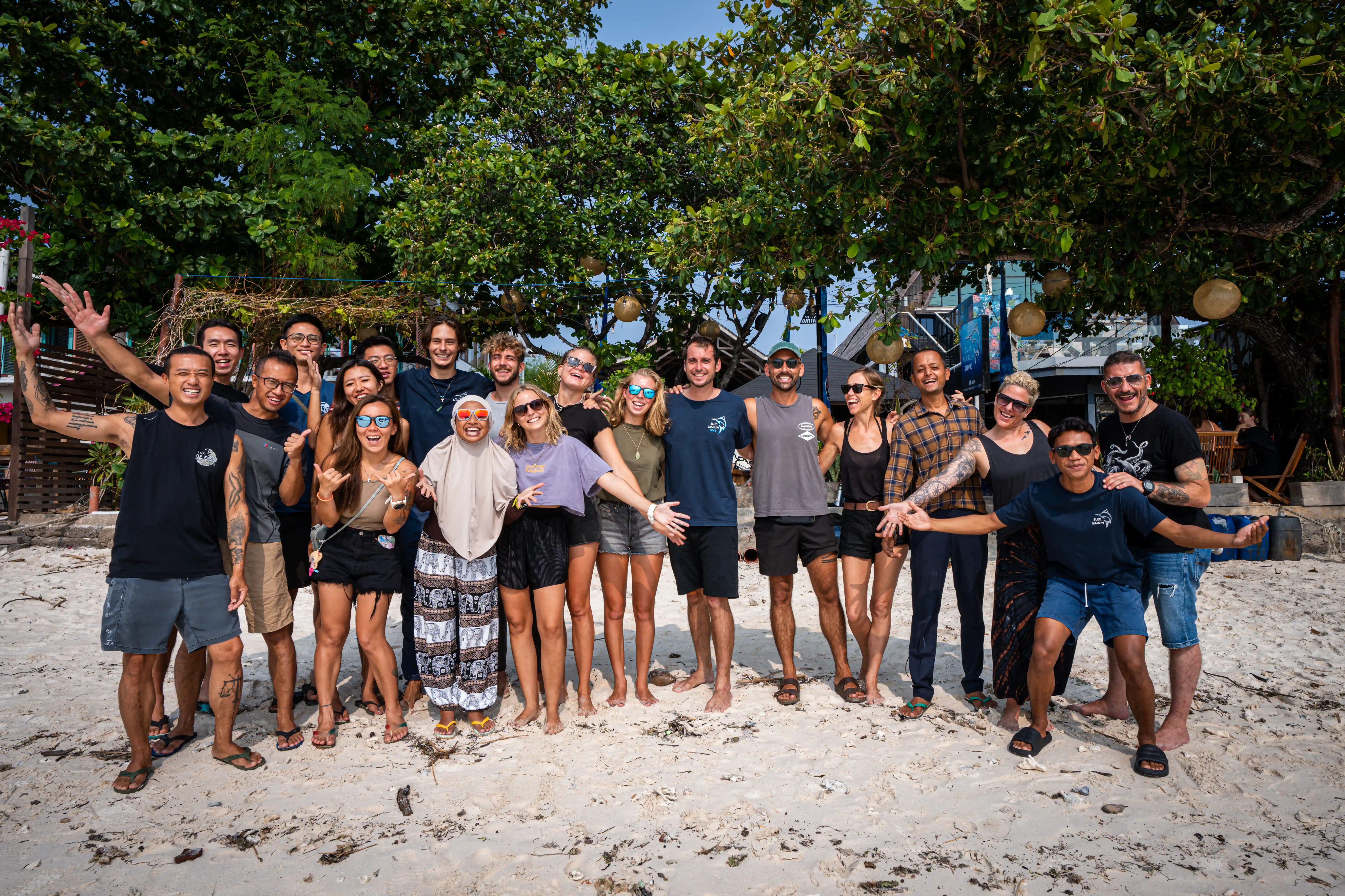 Large group of diverse scuba divers posing together on a sandy beach under tropical trees.
