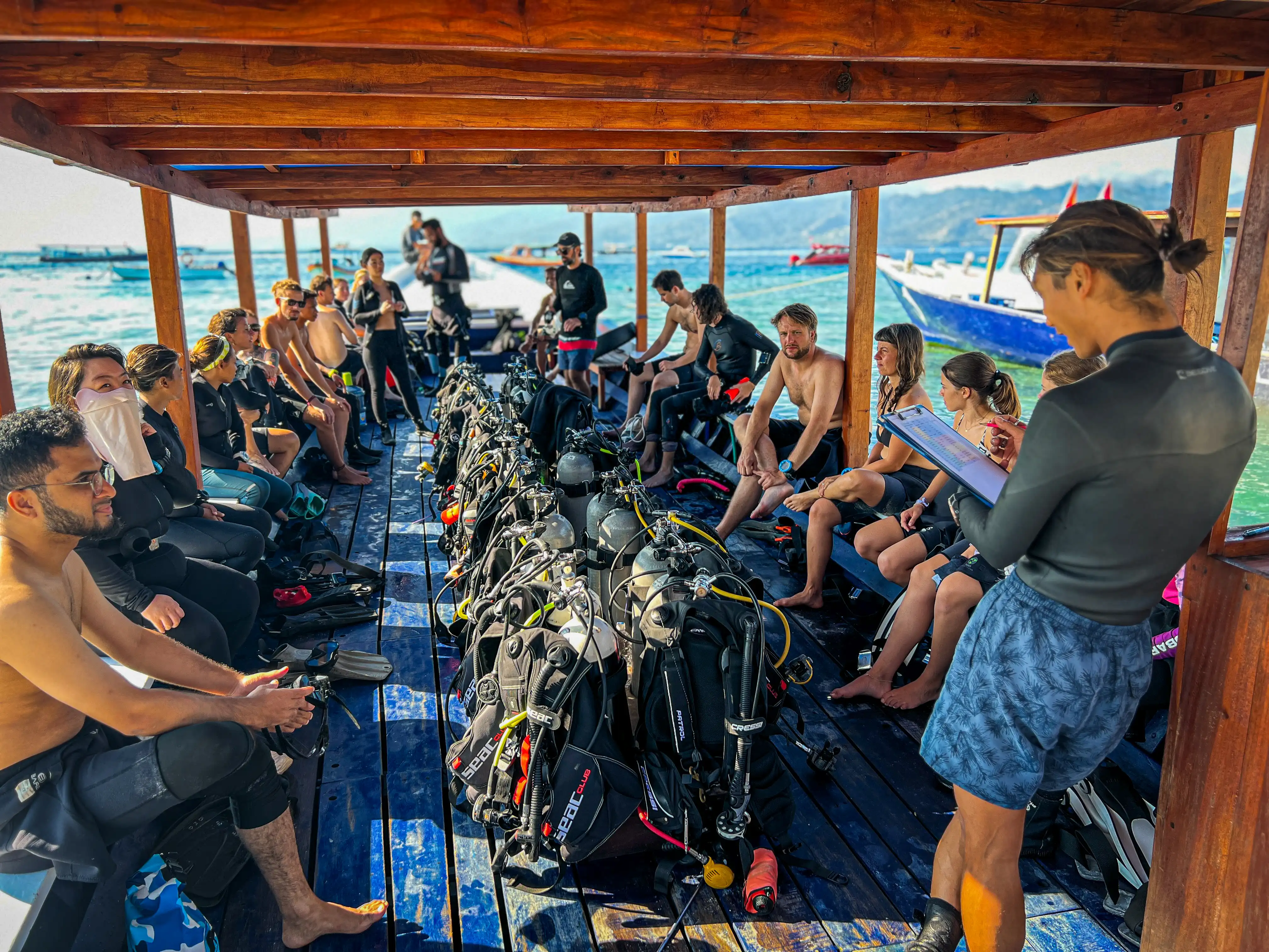 Scuba diving instructor holding a clipboard while briefing divers on a wooden boat.