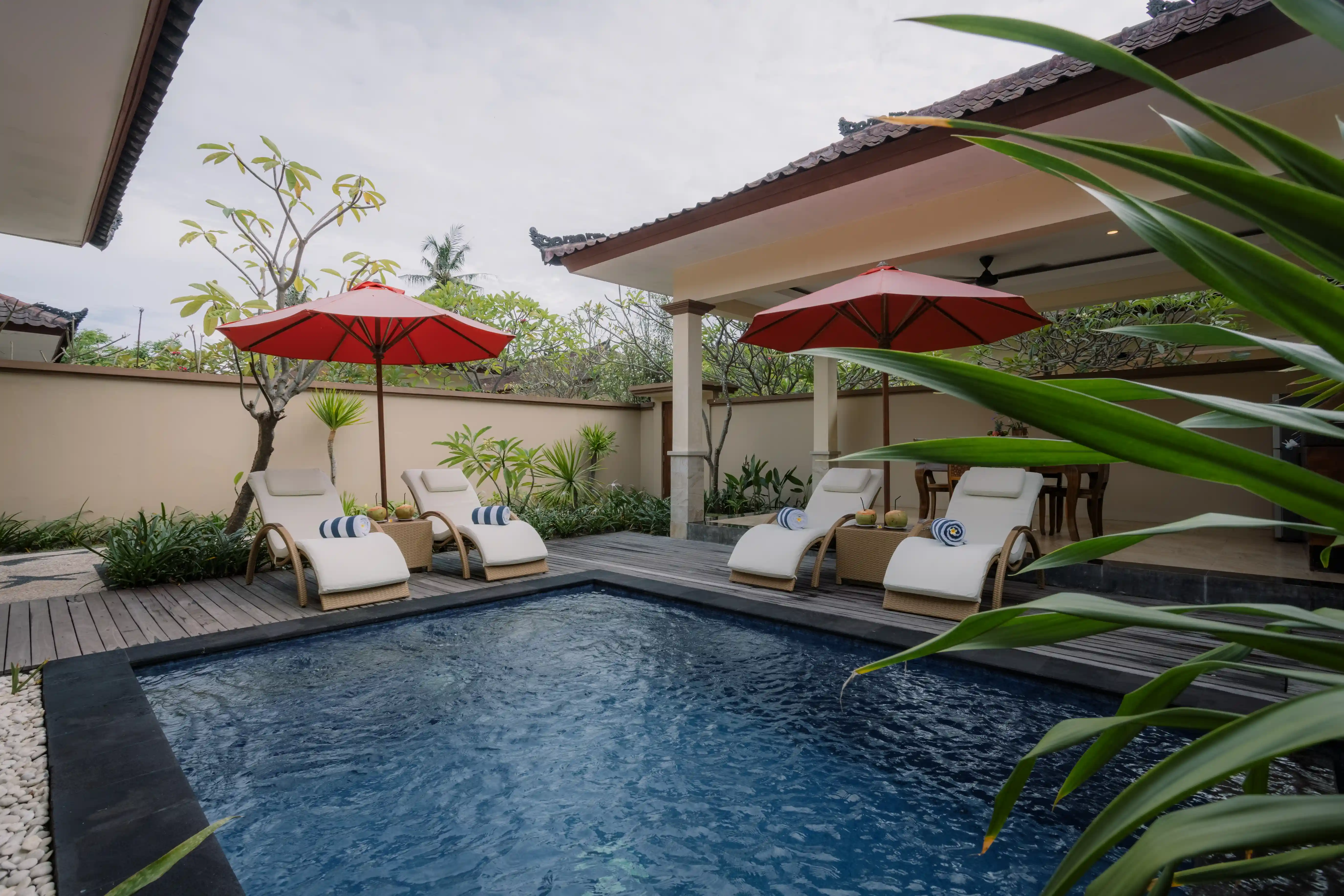 Private outdoor villa pool surrounded by a wooden deck, white sun loungers, red patio umbrellas, and lush greenery.