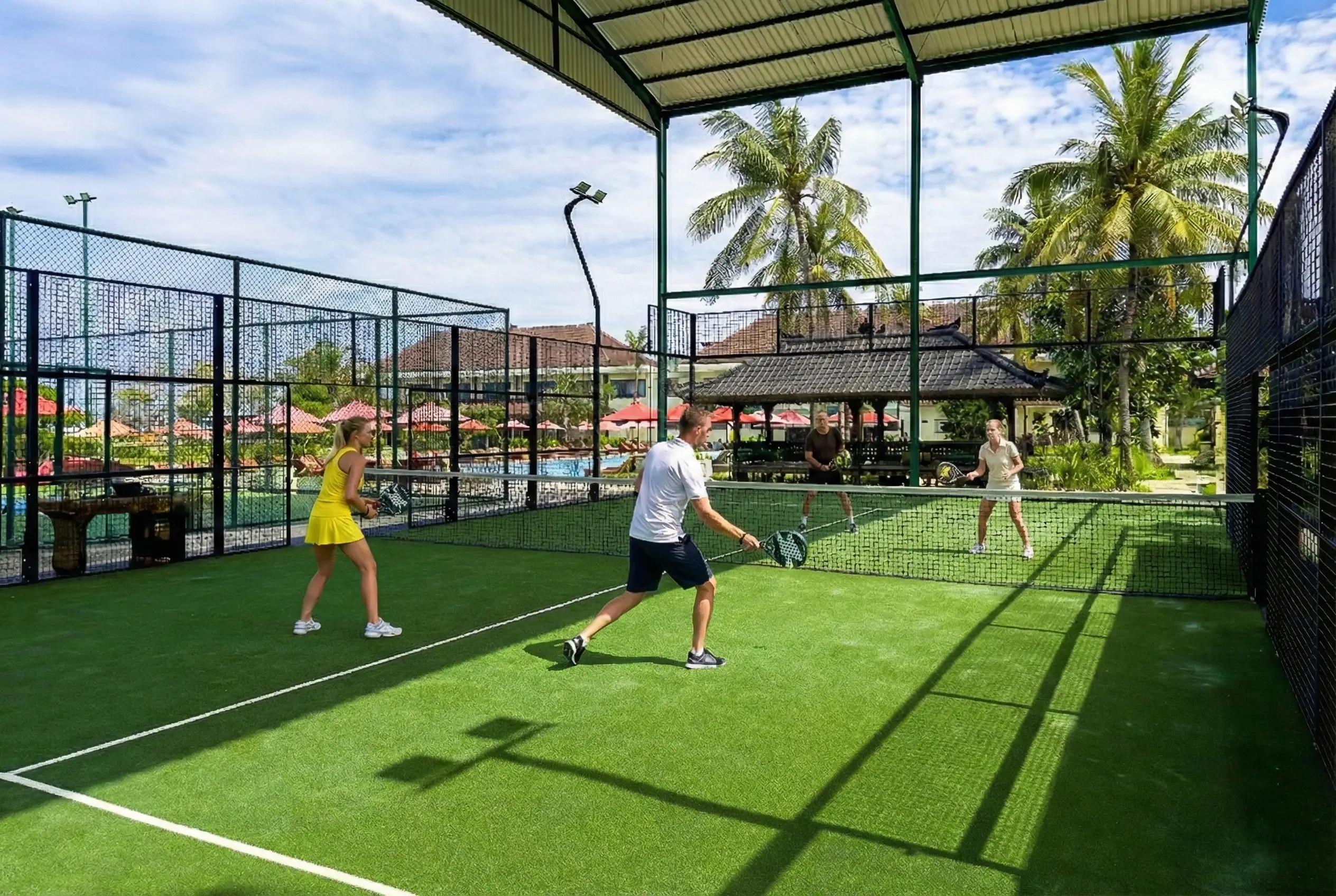 Four people playing padel tennis on a covered green turf court surrounded by palm trees and resort buildings.