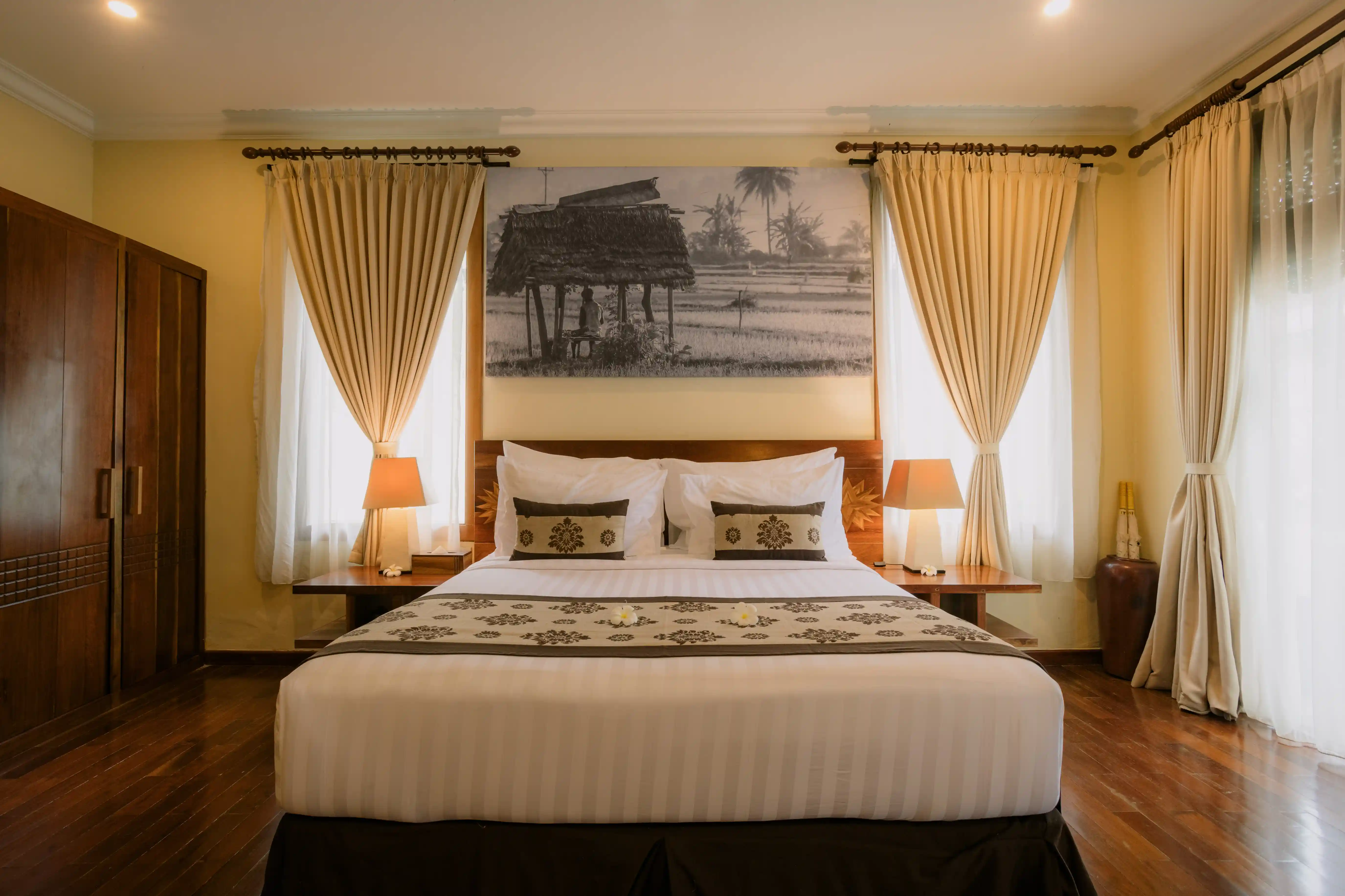 Warmly lit hotel bedroom featuring a king bed with patterned accents, wooden furniture, and warm neutral curtains.
