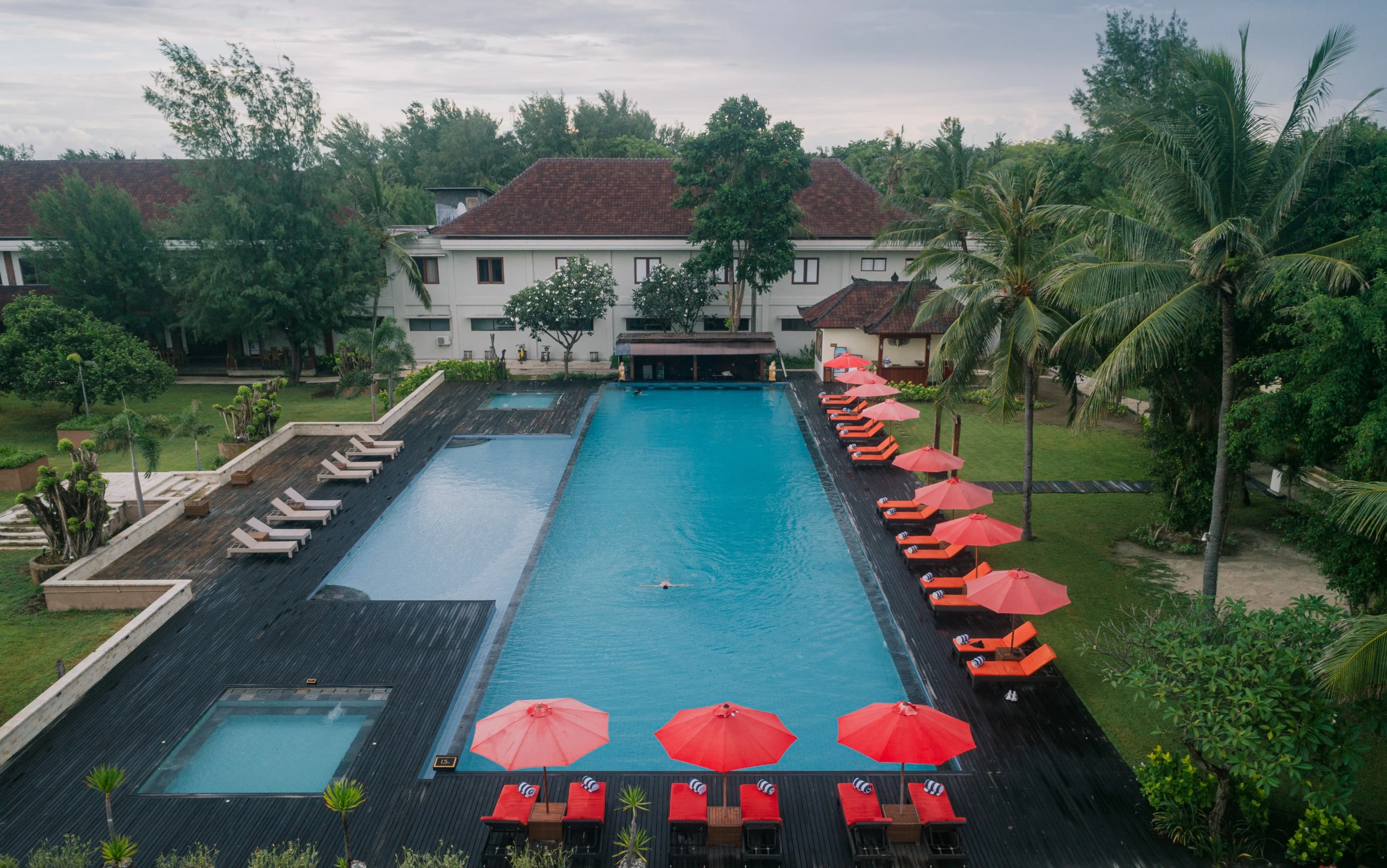 High-angle view of a large rectangular resort swimming pool lined with bright red sun umbrellas and wooden lounge chairs.