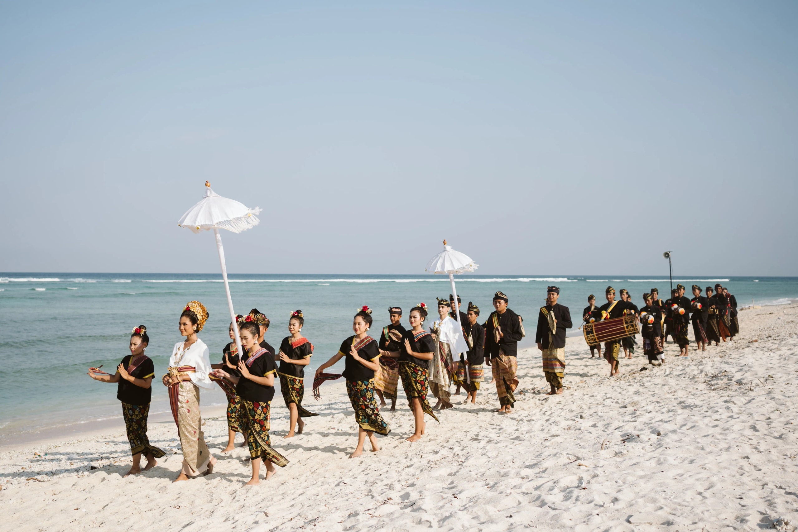 A group of dancers and musicians in traditional Indonesian attire performing a procession along a sandy beach.
