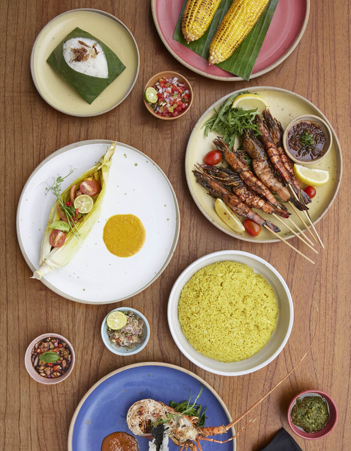 Flat-lay spread of grilled seafood, rice, corn, and traditional Indonesian dipping sauces.