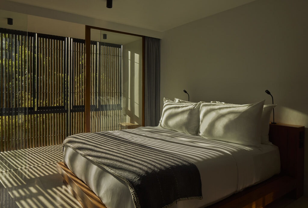 Sunlit villa bedroom with slatted wooden blinds casting shadows over the bed.