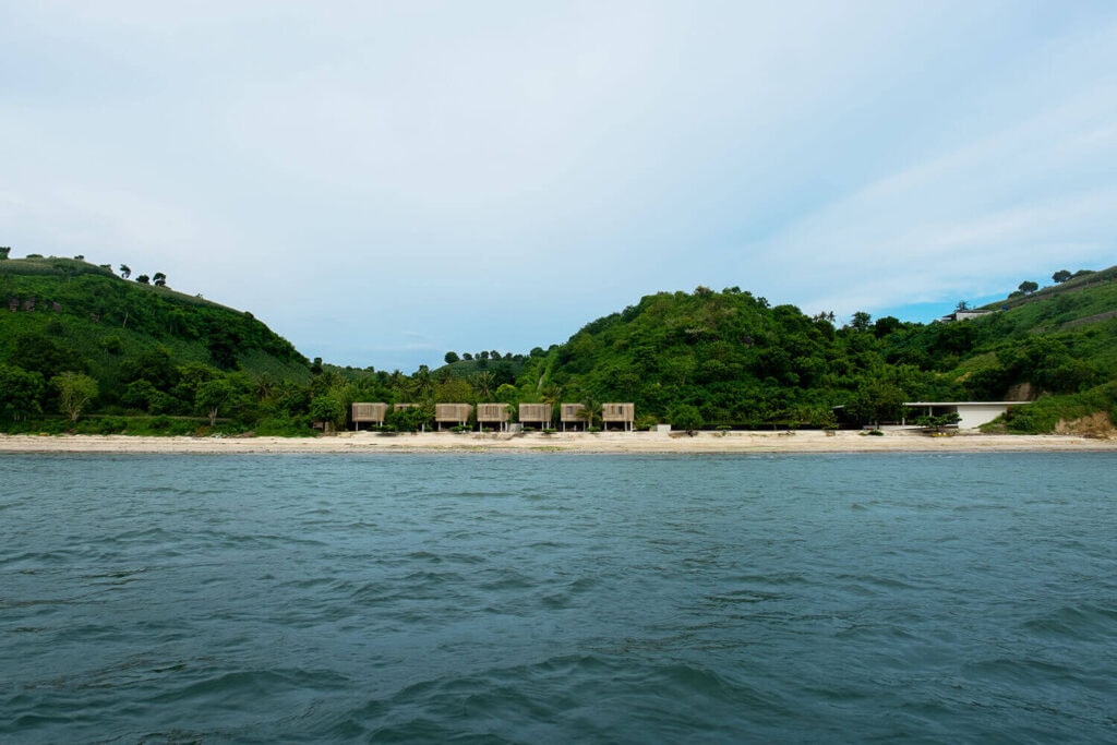 Distant ocean view of beachfront wooden villas nestled against green hills.