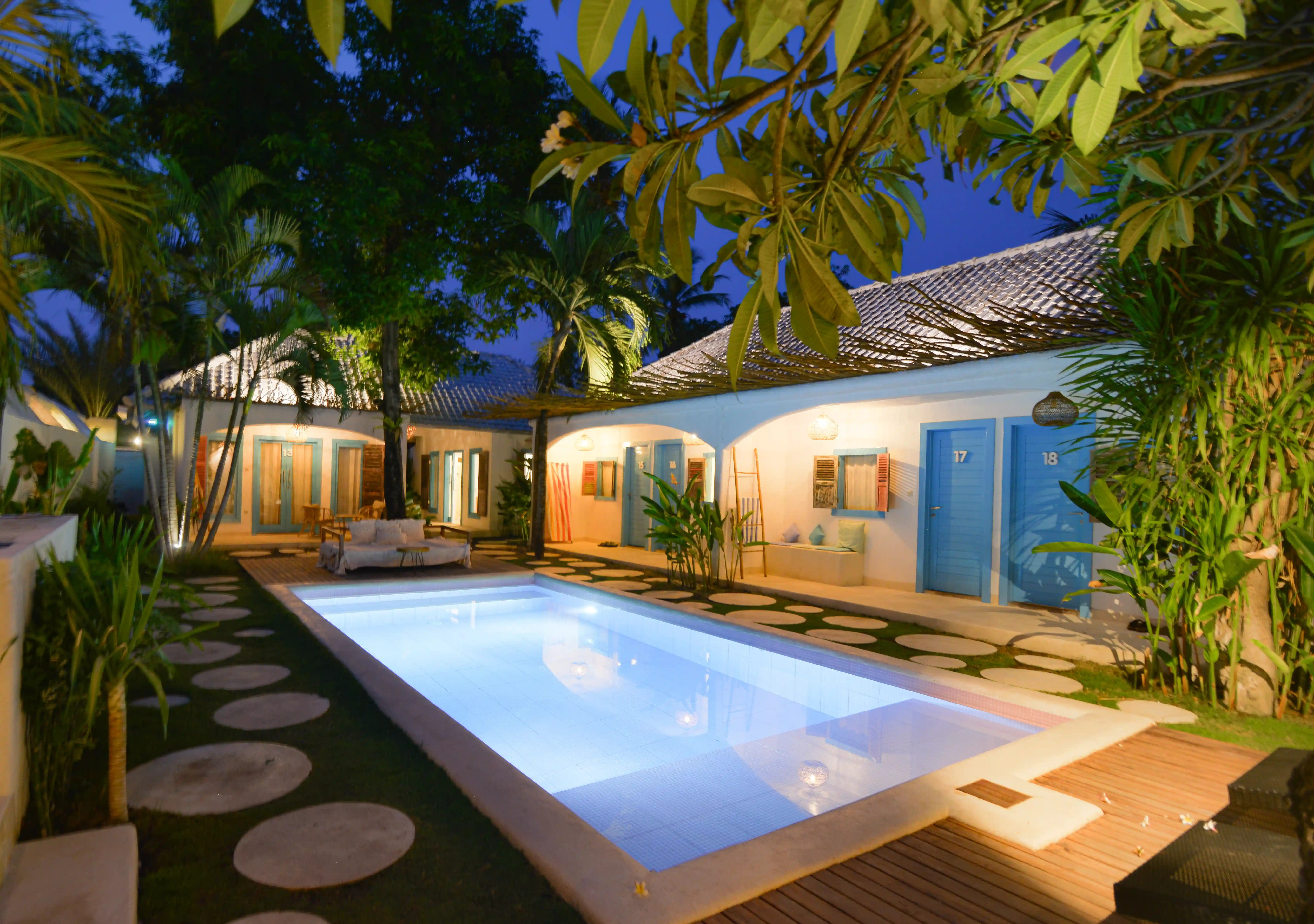 Piscine illuminée de nuit au milieu de bungalows blancs aux portes bleues dans un cadre tropical.
