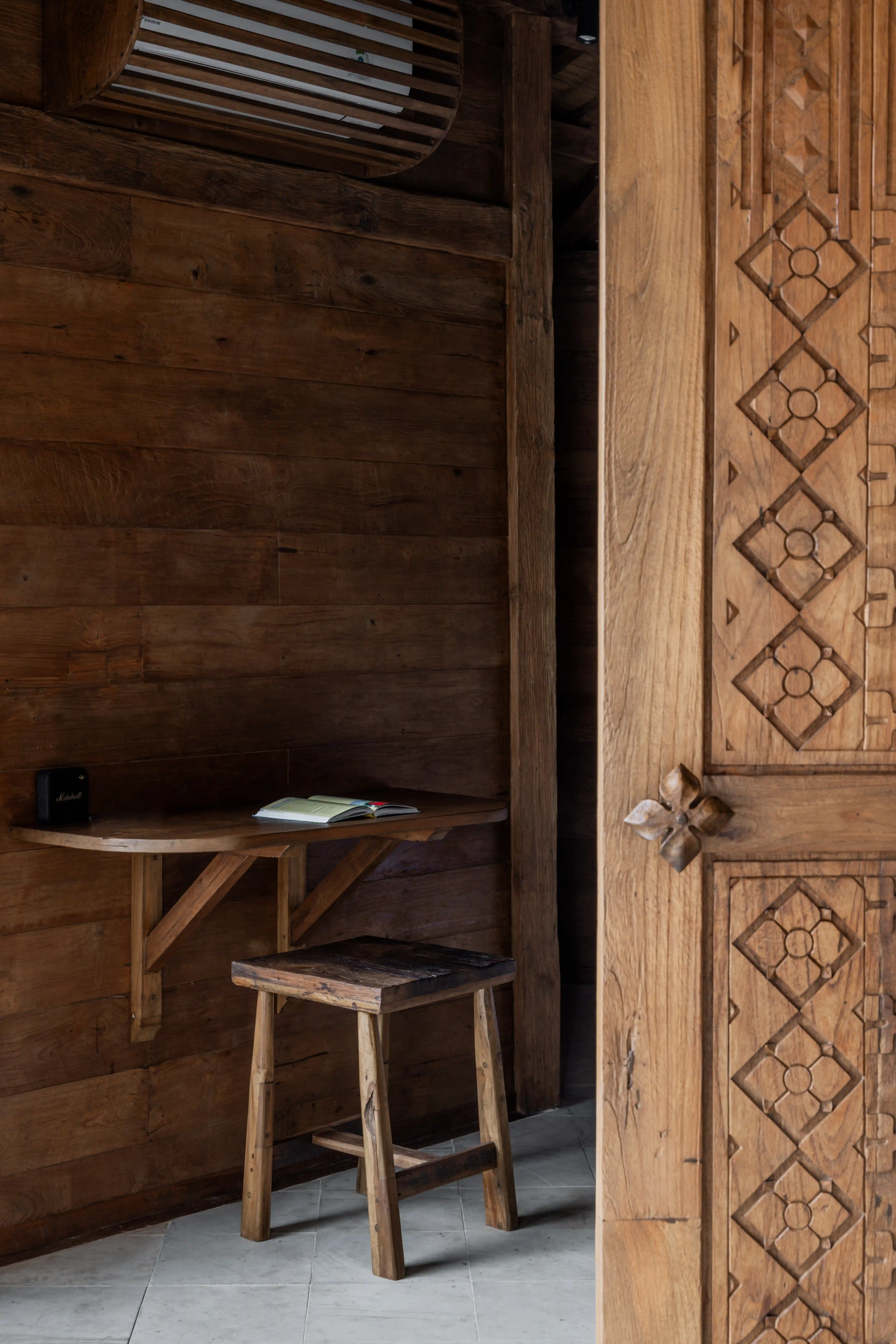 A small wooden writing desk and rustic stool in a room with warm wooden walls and a carved wooden door.