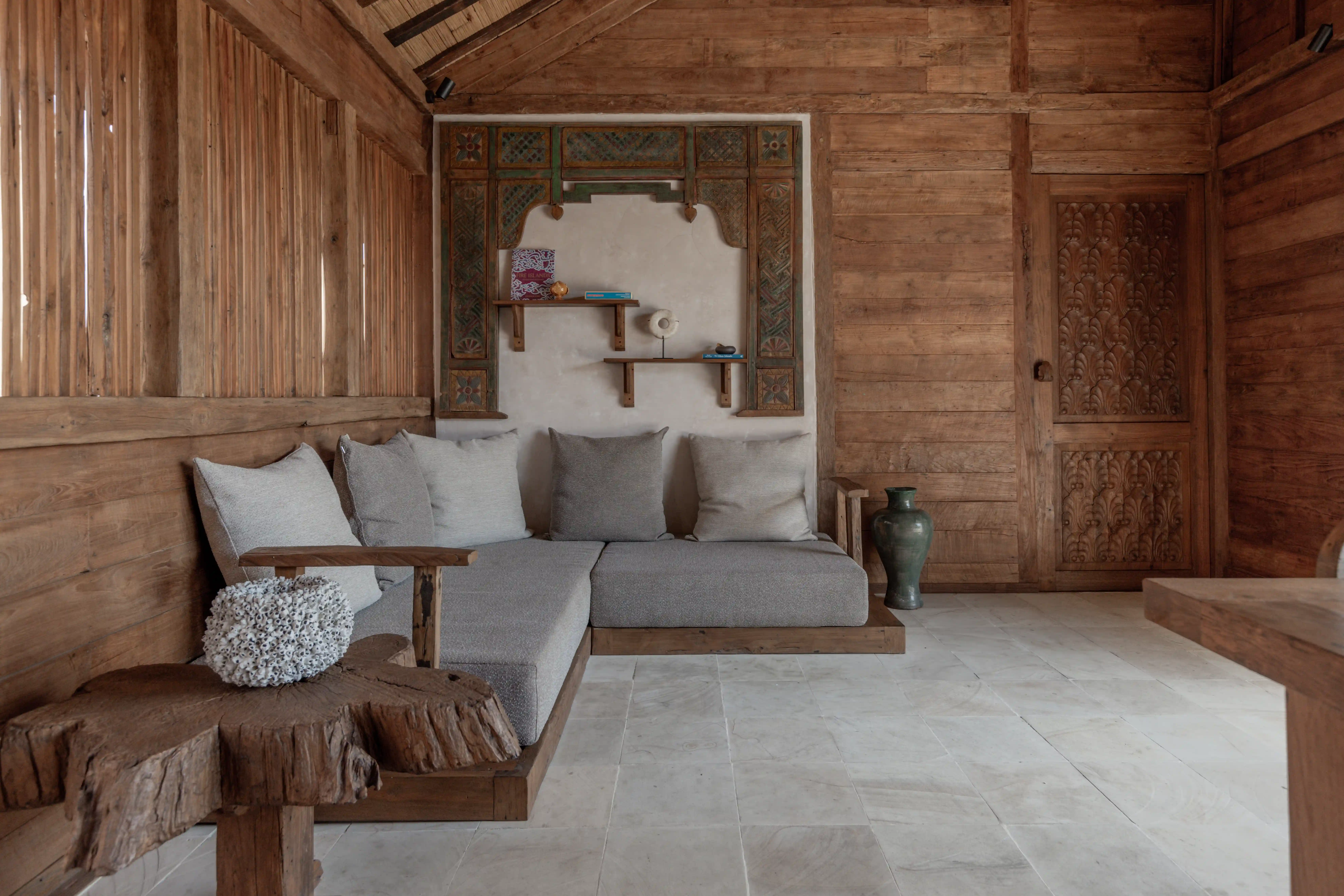 Cozy indoor lounge area with a grey L-shaped sofa, carved wooden wall accents, and a natural log coffee table.