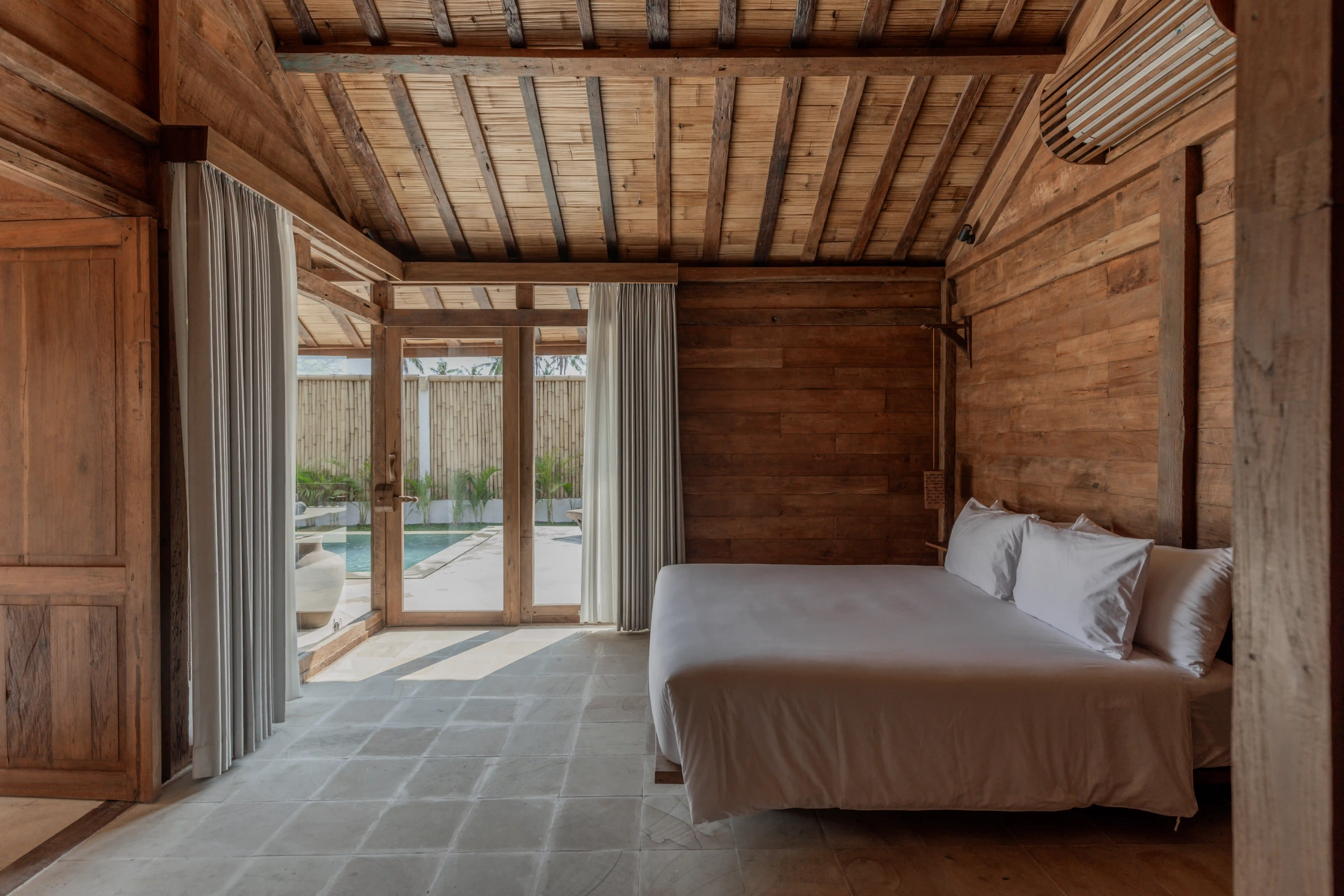 Bedroom with a king-size bed, exposed wooden walls, woven bamboo ceiling, and glass doors leading to the pool.