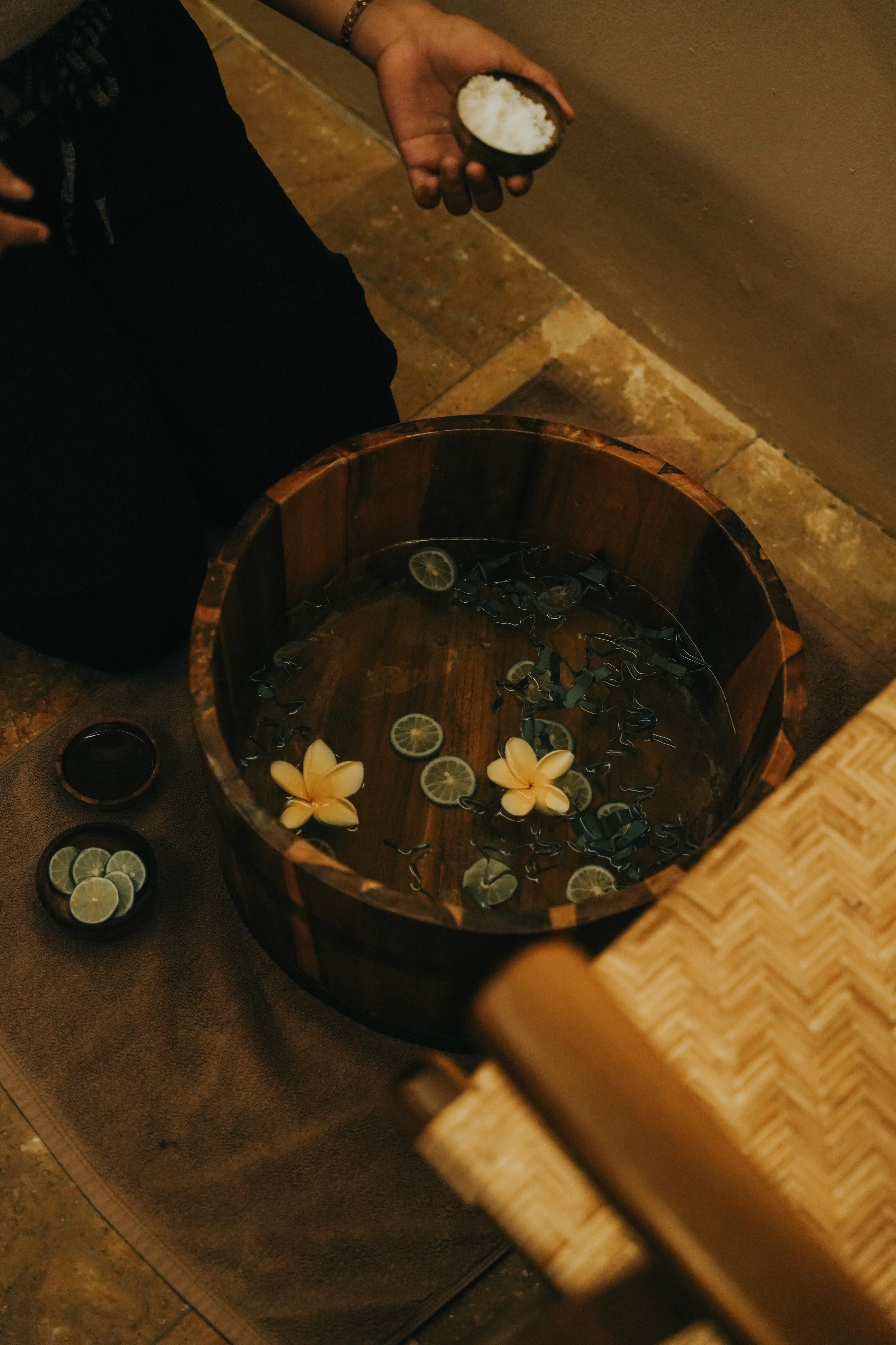 igh-angle view of a wooden foot bath basin filled with water, lime slices, and yellow frangipani flowers, with a therapist holding a small bowl of bath salts above.