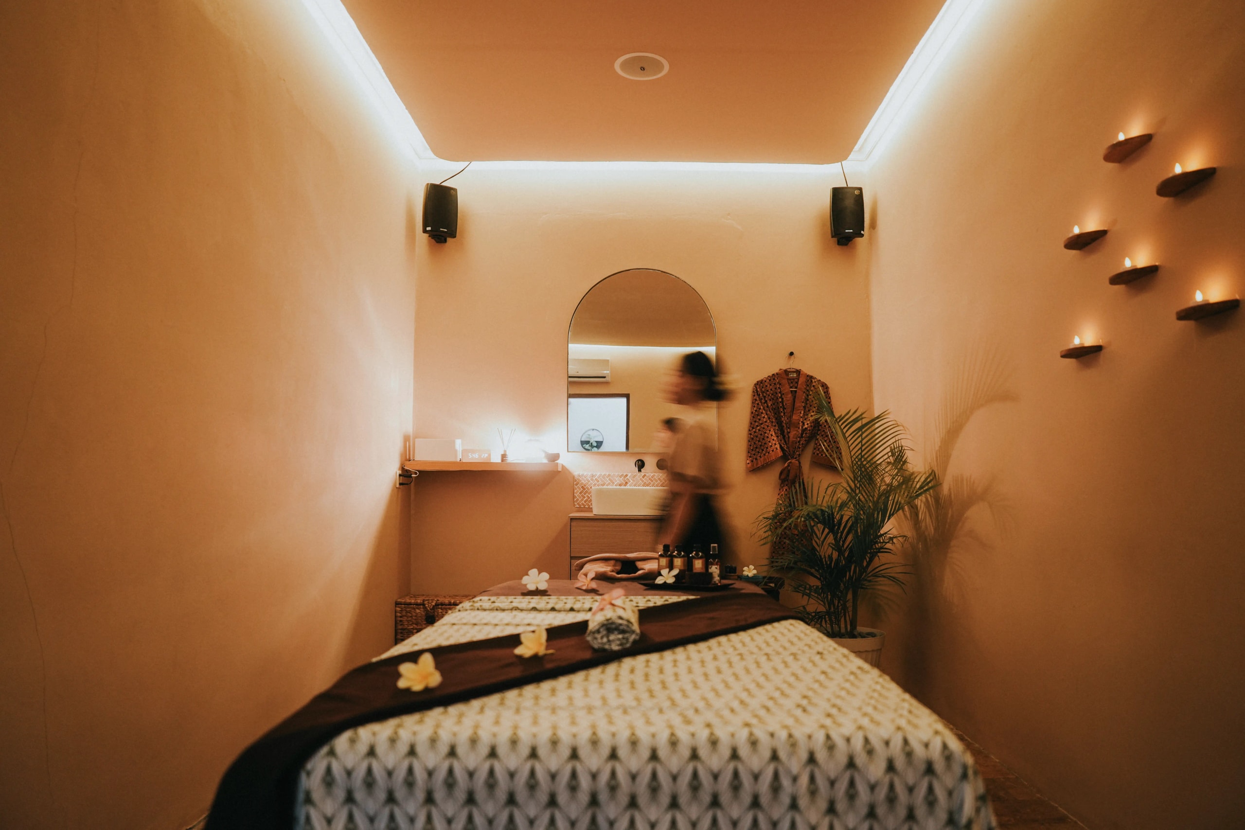 Warmly lit single treatment room featuring a prepared massage table with patterned linen, wall-mounted candles, and a vanity in the background.